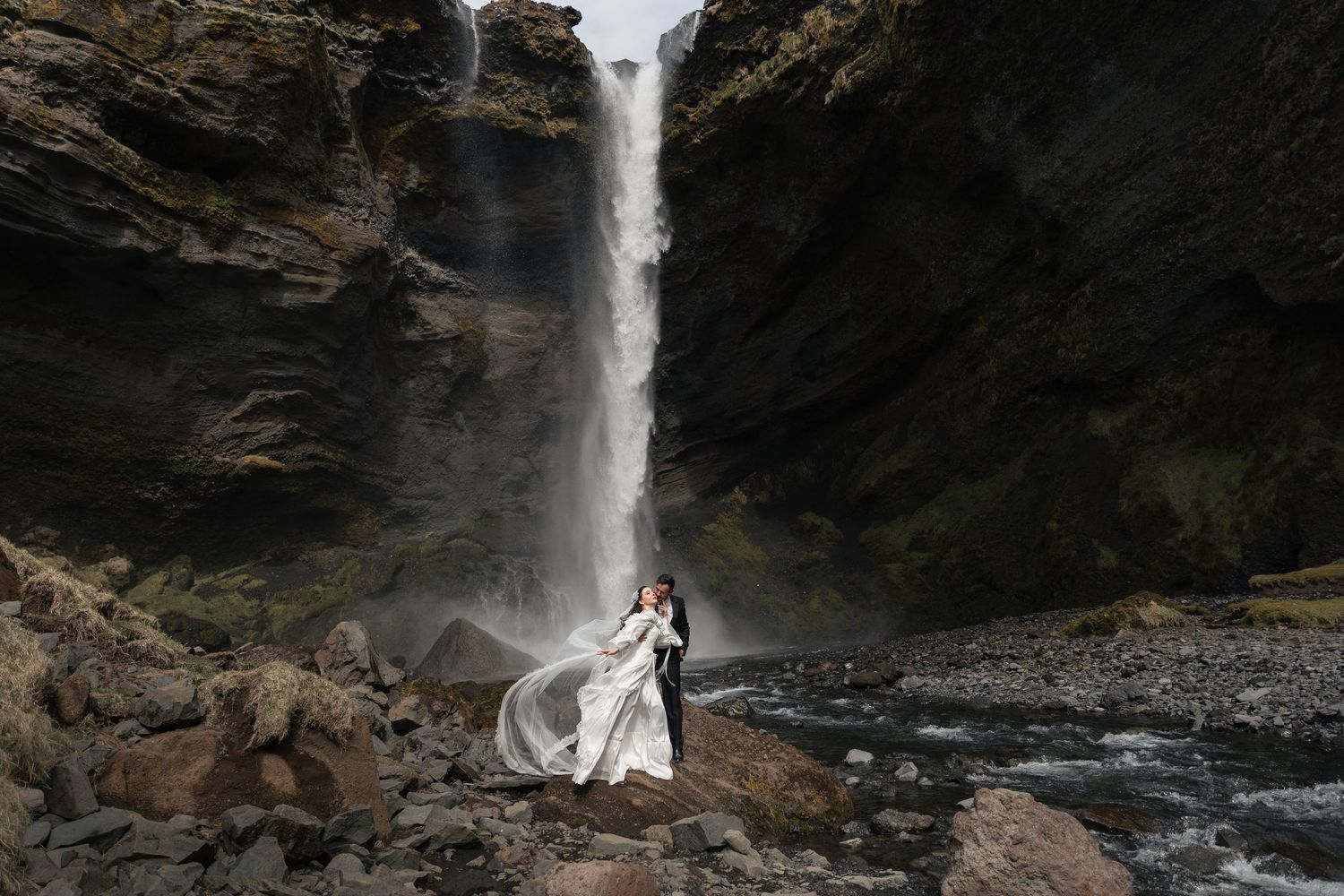 Couple embracing in wedding attire before a dramatic waterfall cascading between rocky cliffs.
