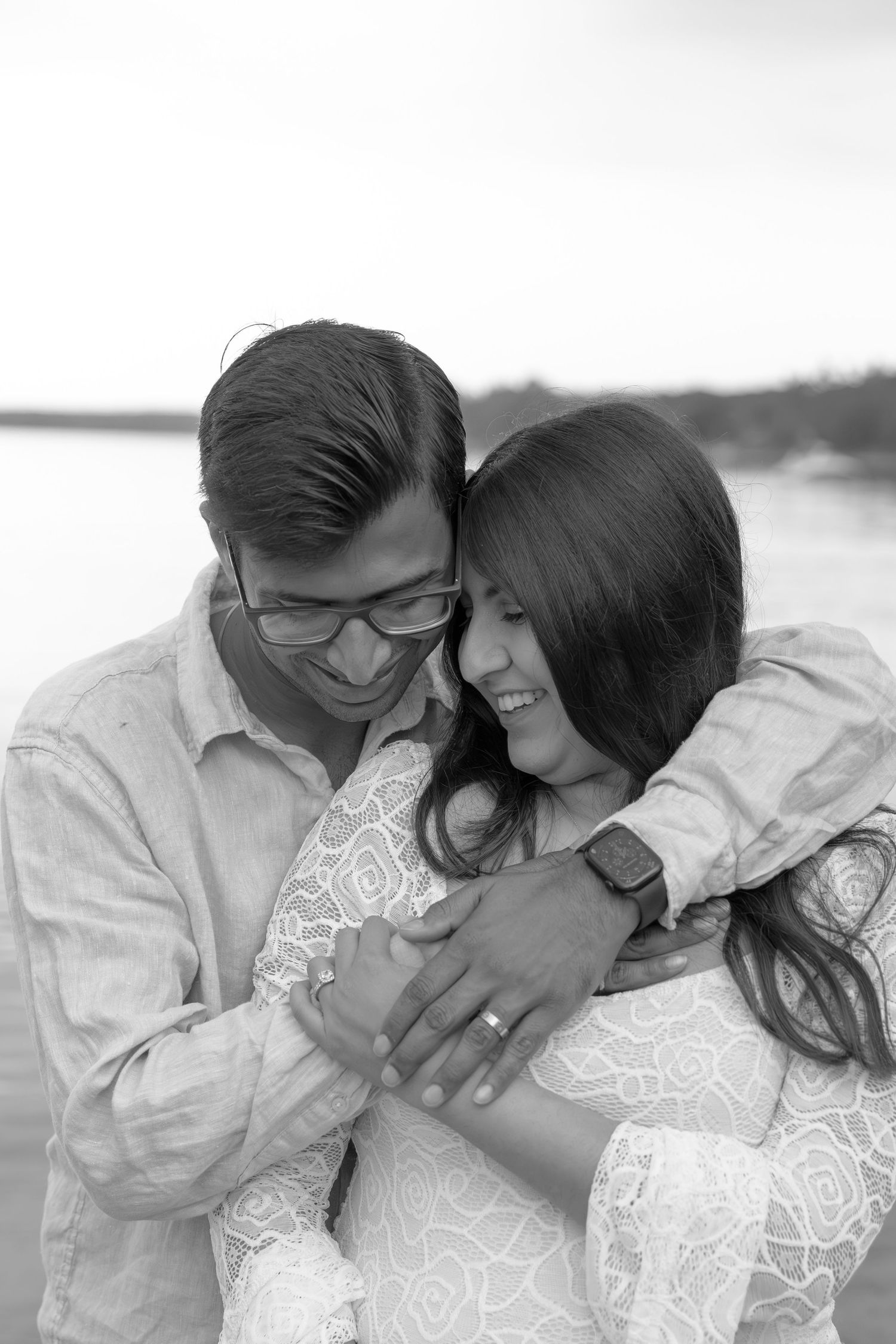 A romantic black and white portrait of a couple embracing and laughing together by the water.