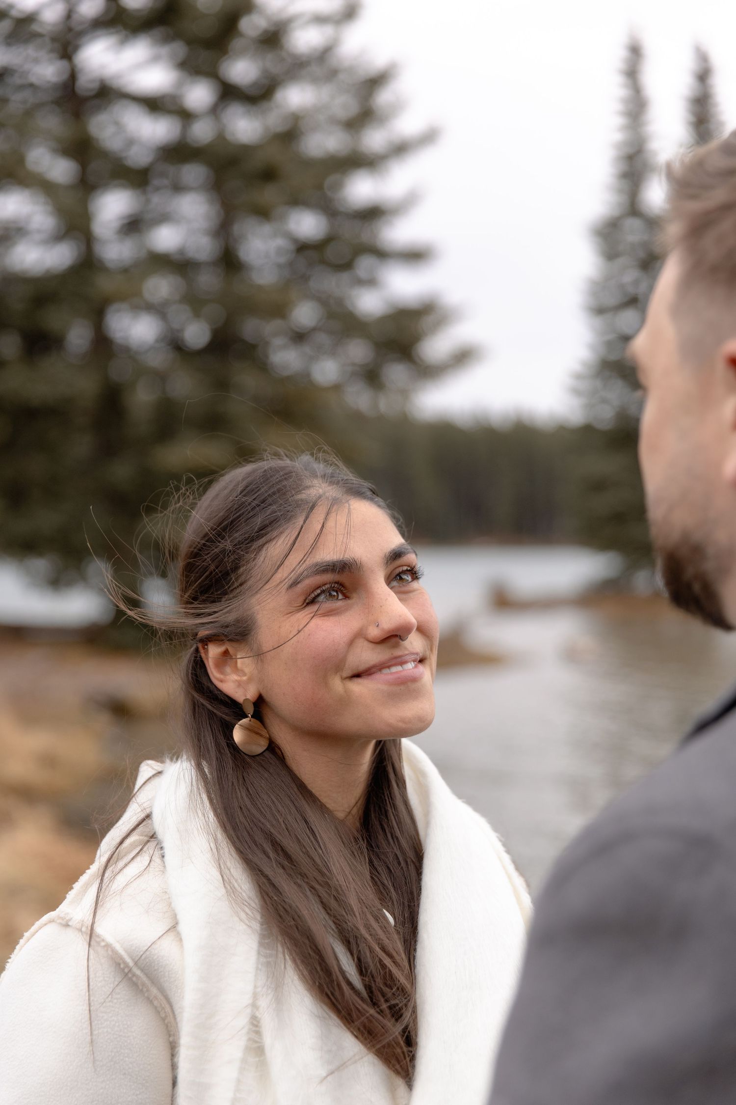 A couple shares an intimate moment in a winter forest setting with evergreen trees and snow in the background.