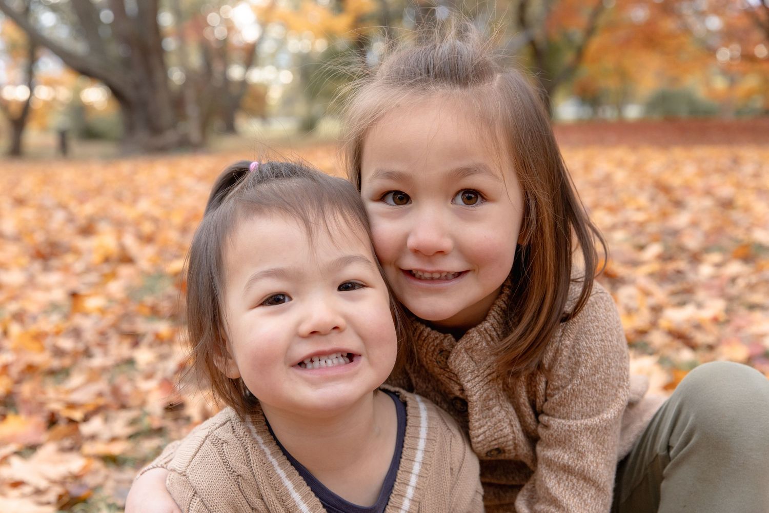 Two children smile together while sitting on autumn leaves in a park during fall season.