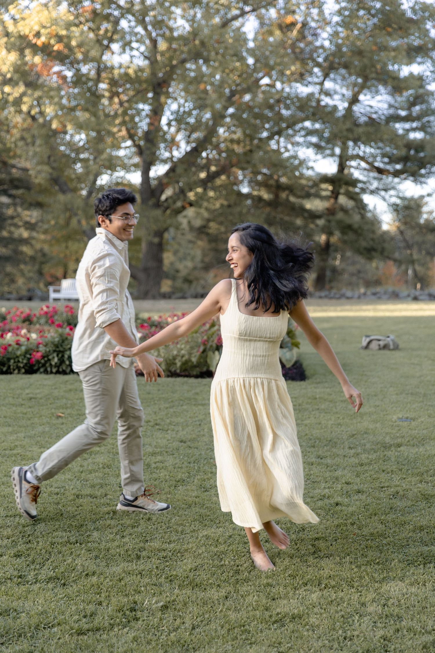 A couple in white outfits playfully jumps and dances together on a grassy field during a sunny day.