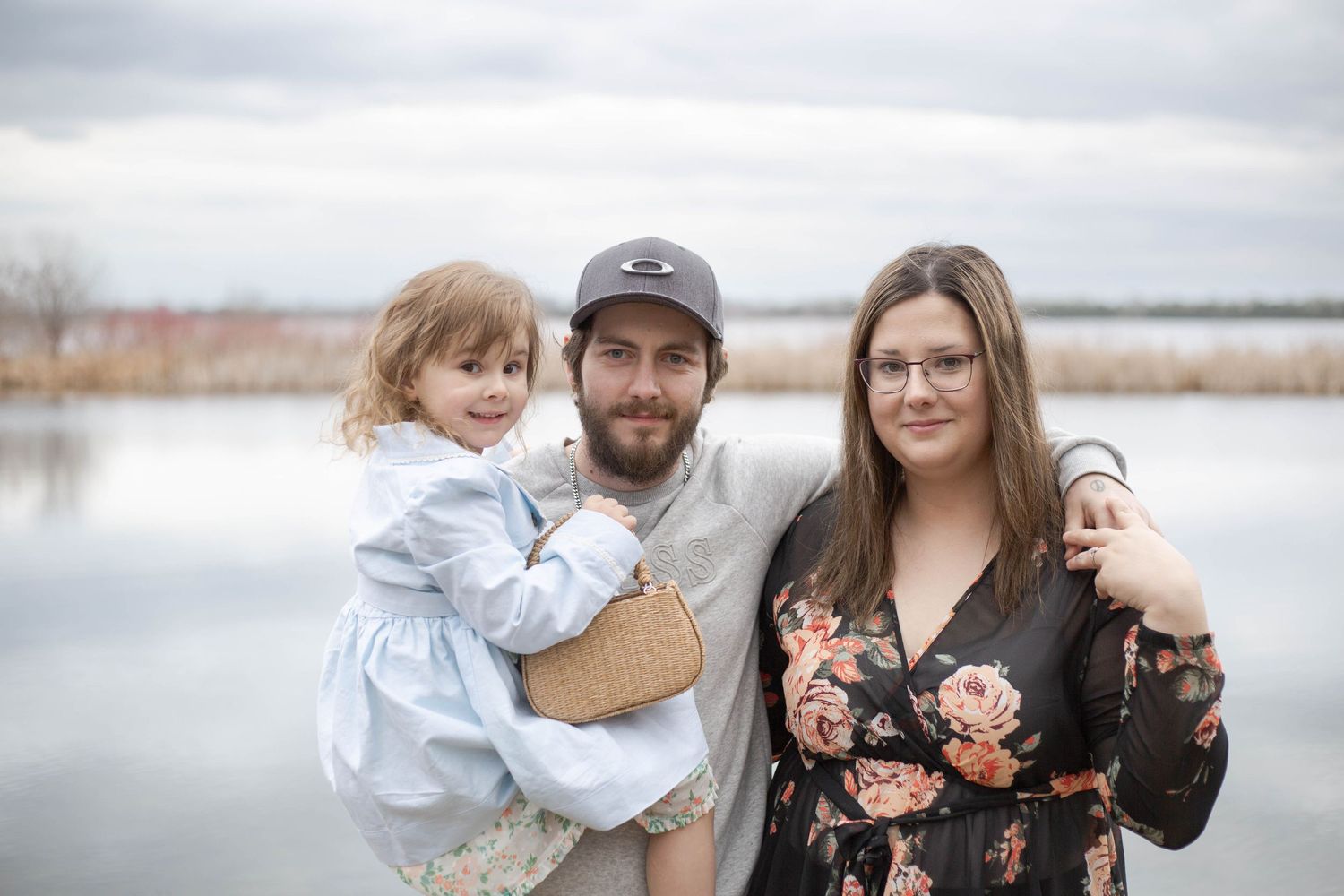 A family poses together by a serene lake on an overcast day with calm water and bare trees in the background.