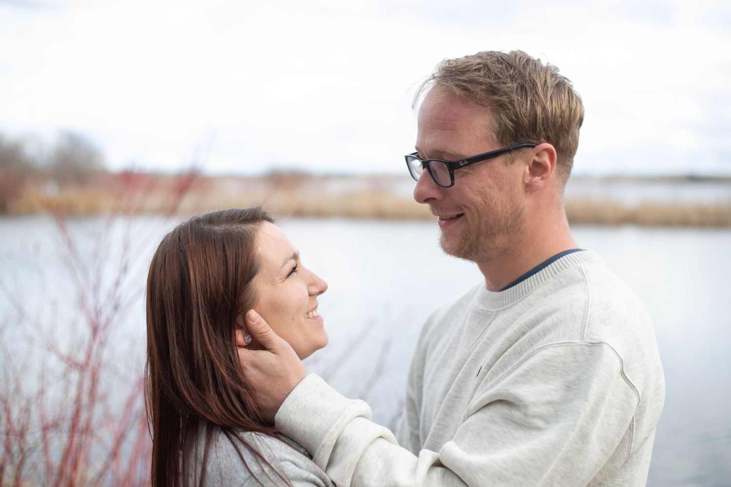A couple wearing cozy sweaters share a loving moment while standing by a lake on a winter day.