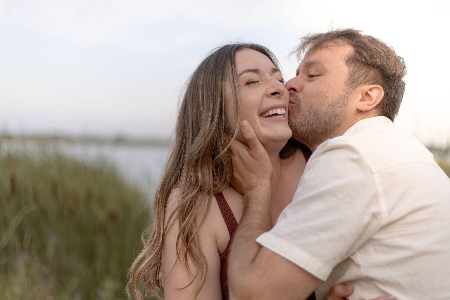 A romantic couple embraces in a sunlit meadow wearing casual white clothing.
