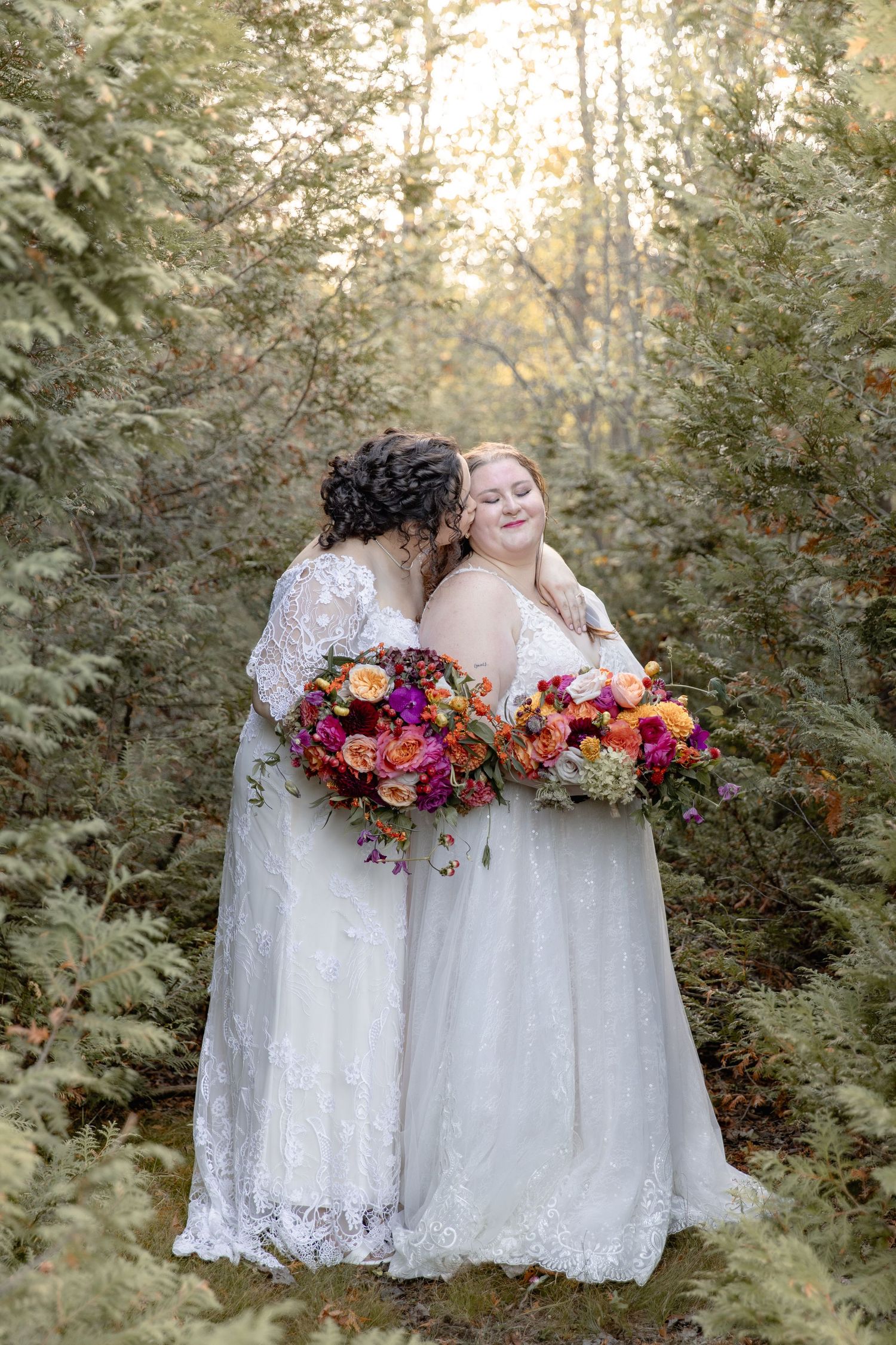 Two people in white lace dresses share a joyful moment with vibrant autumn florals in a forest setting.