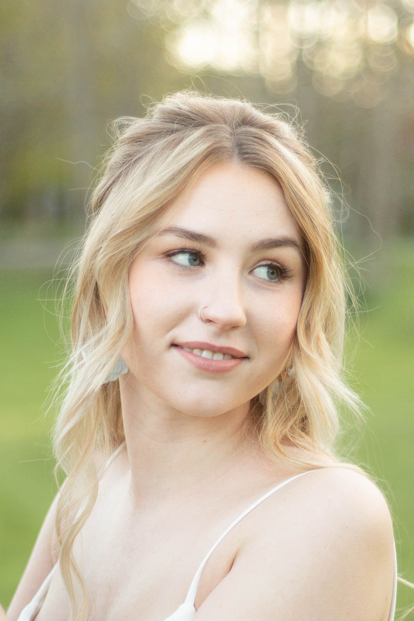 Romantic outdoor portrait shows a woman with blonde wavy hair wearing a light tank top against a soft natural background.