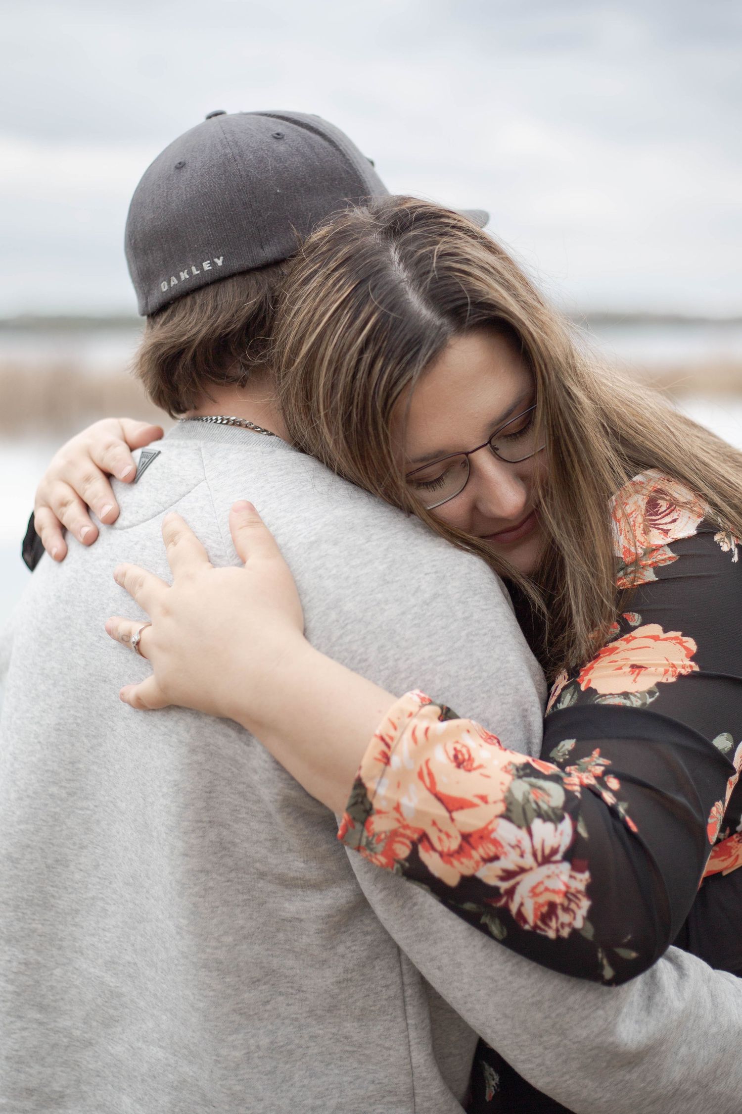 A couple embraces in a tender moment at the beach, with one wearing a floral top and the other in a gray sweatshirt.