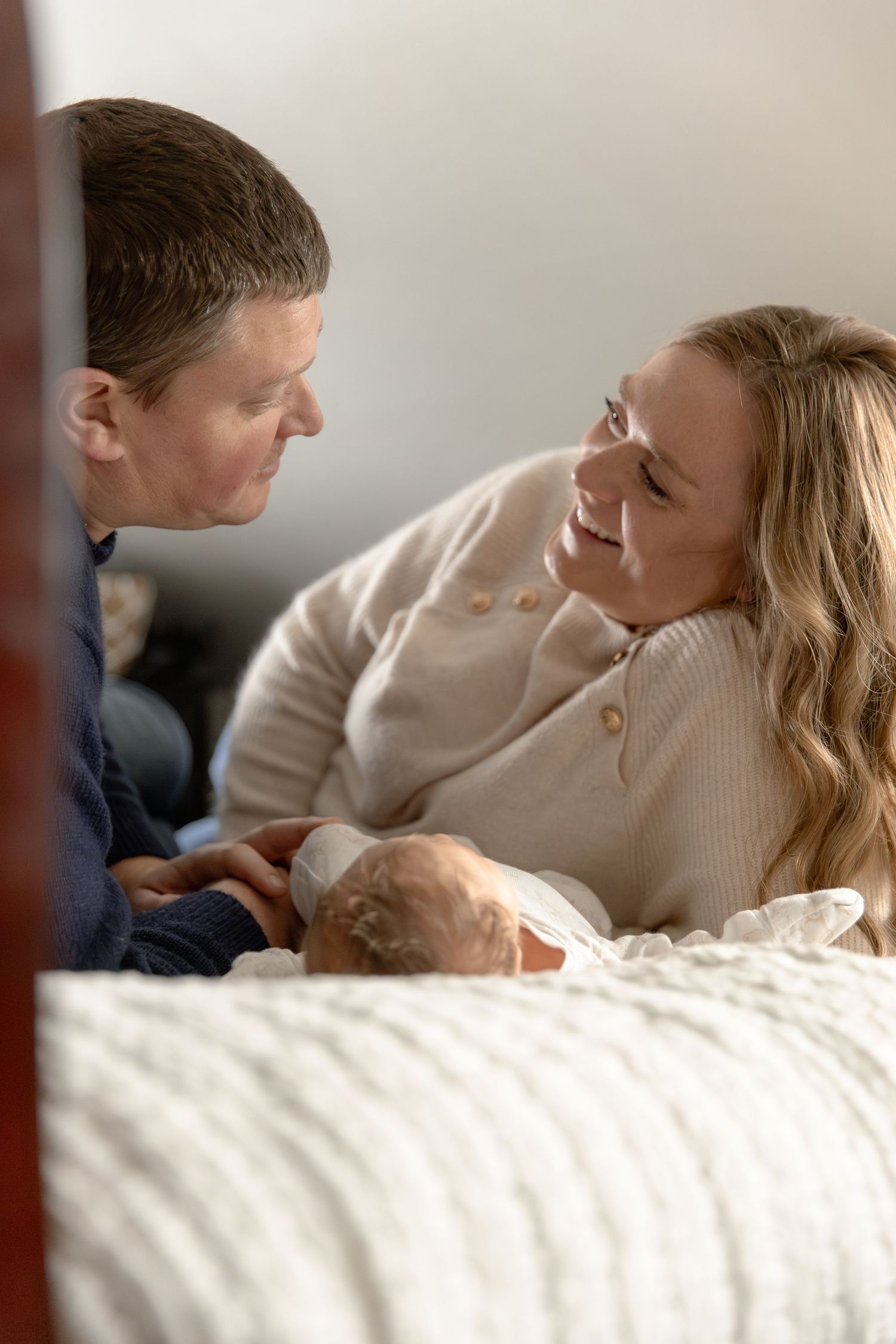 A couple shares an intimate moment while lying together on a white bed, both wearing light colored sweaters.