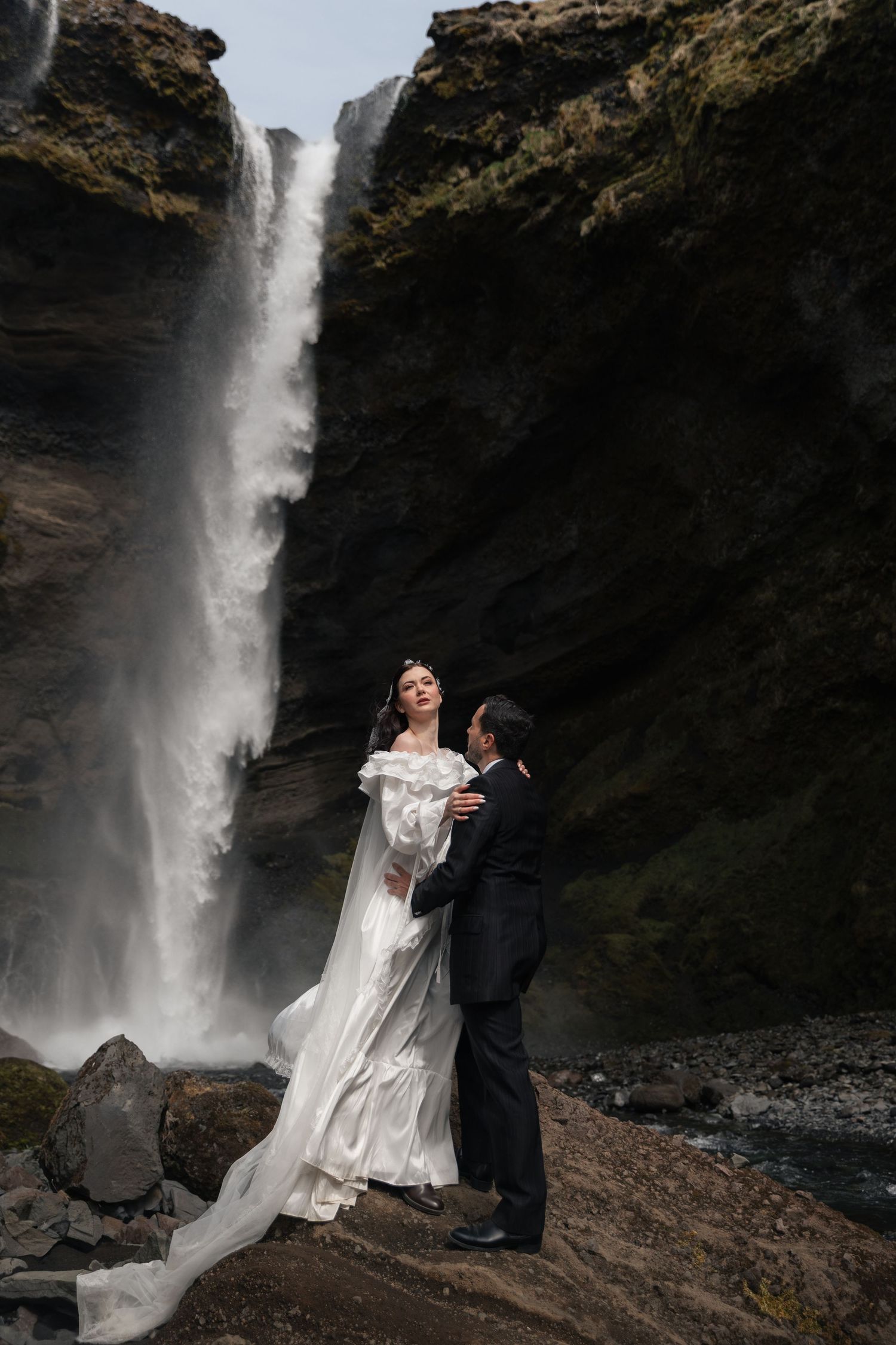 Bride and groom embrace before a dramatic waterfall in Iceland, bride wearing flowing white off-shoulder gown.