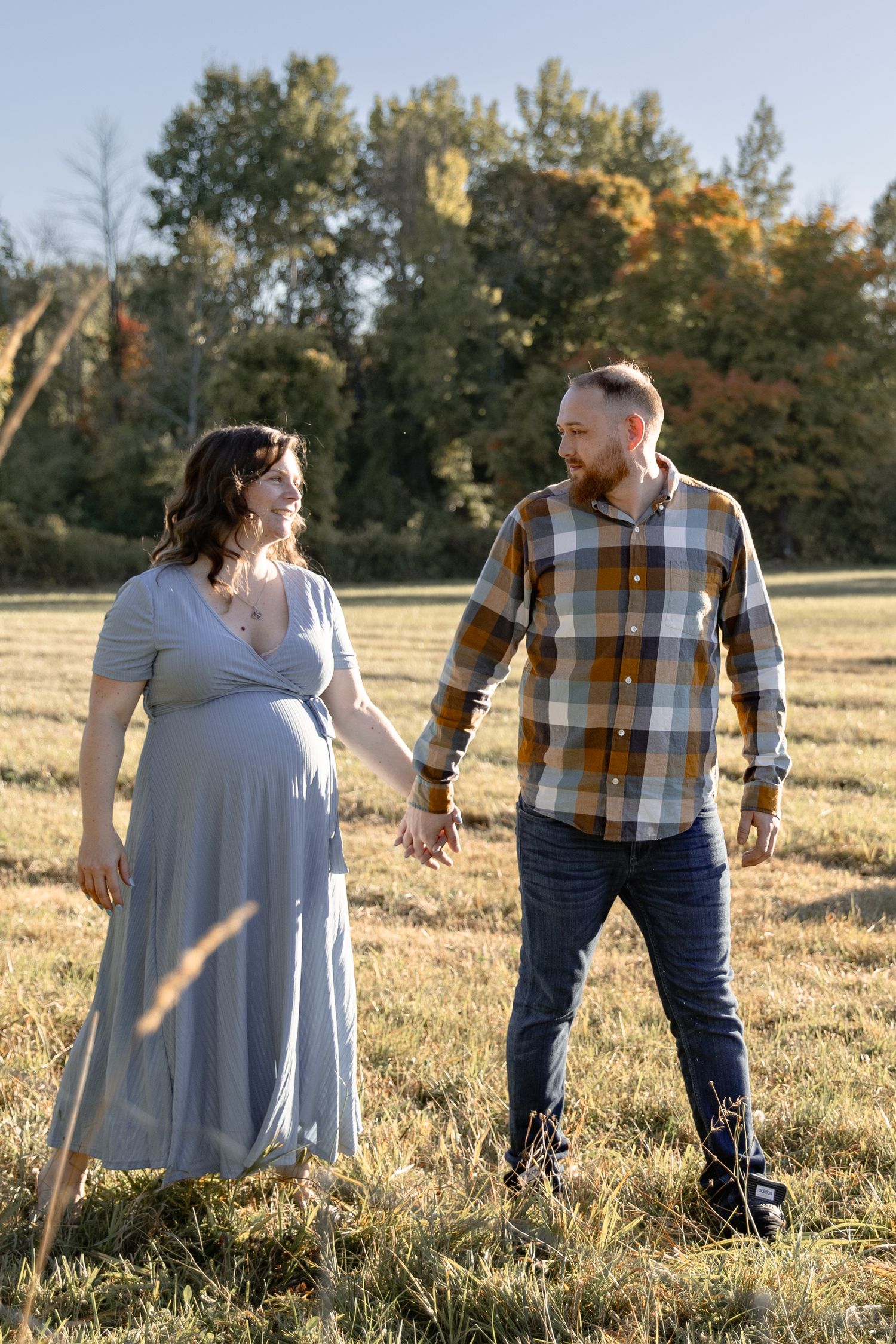 A couple walks hand in hand through a golden autumn field at sunset in casual fall attire.