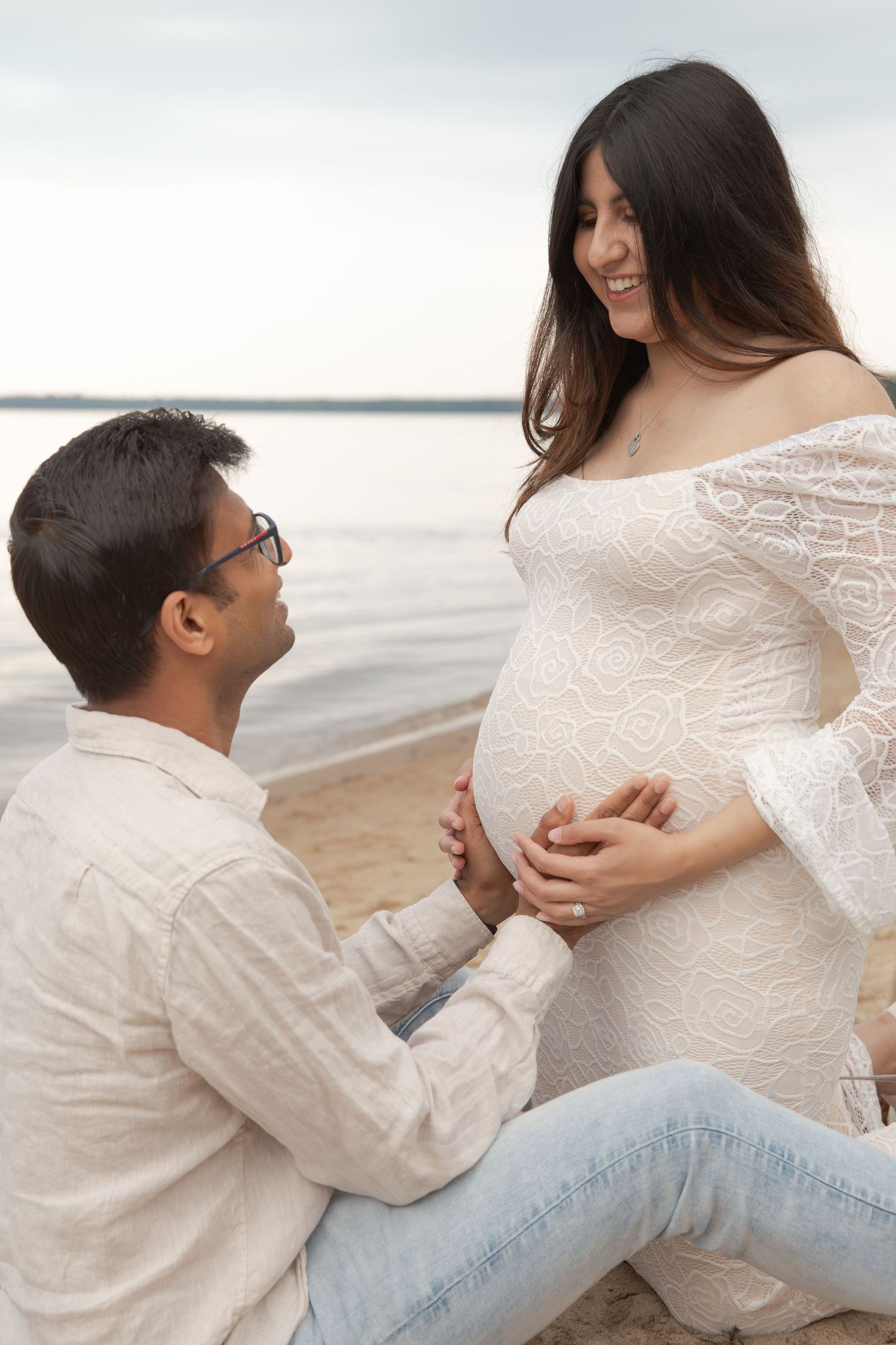 Intimate beachside maternity photoshoot with couple in white attire sharing tender moment.