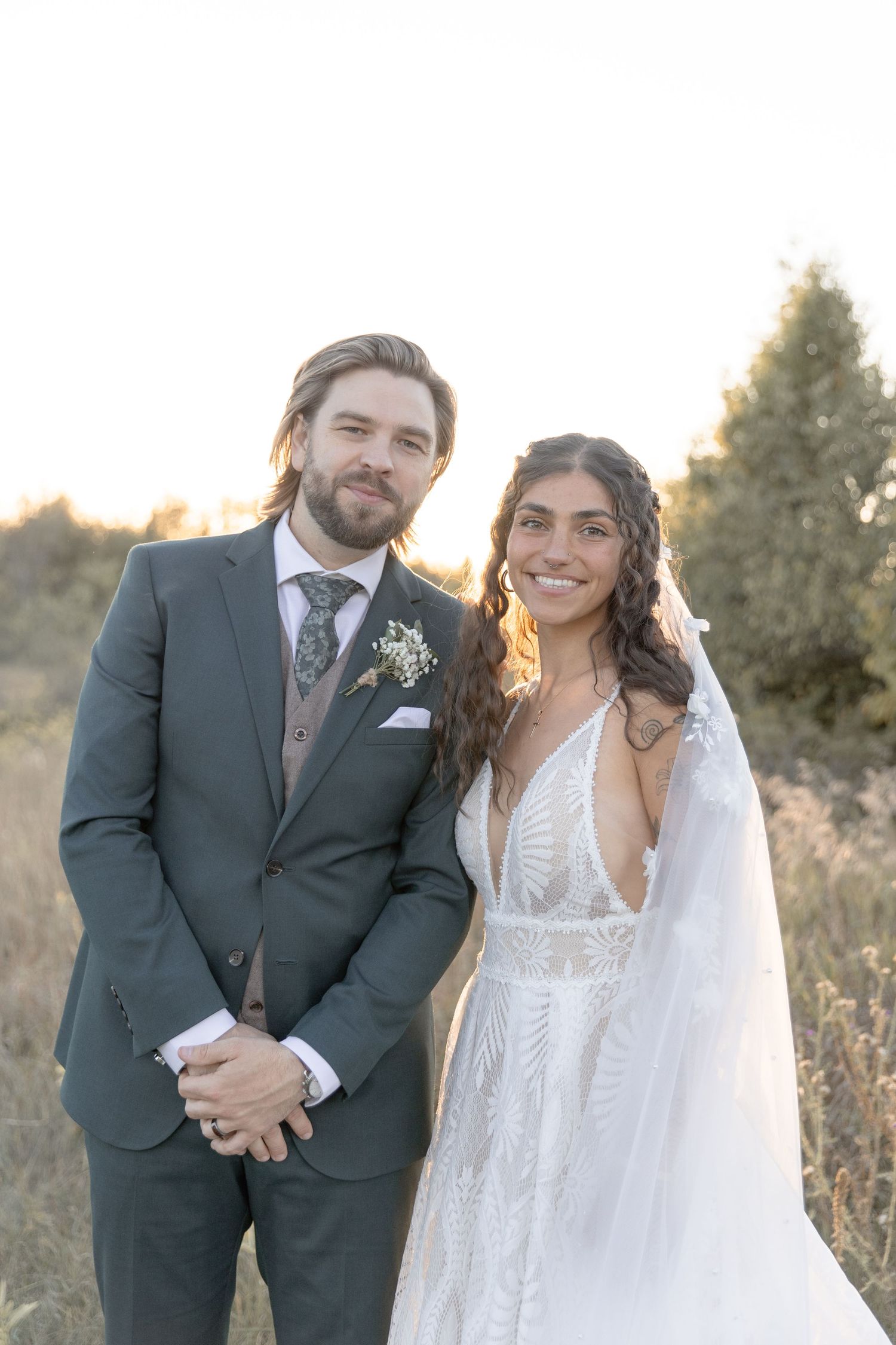 A couple poses together in an outdoor autumn setting wearing formal wedding attire at sunset.