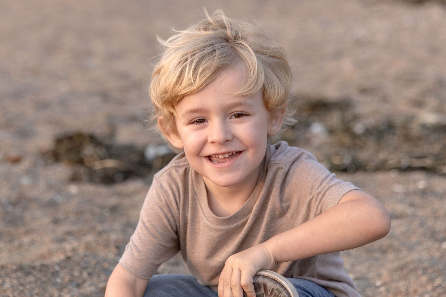 A joyful child with blonde hair smiling at the camera while sitting outdoors.