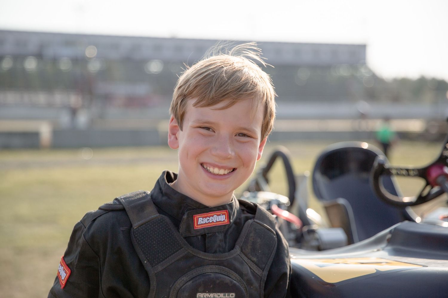 Young race car driver wearing black safety gear smiles while at outdoor racing track on sunny day.