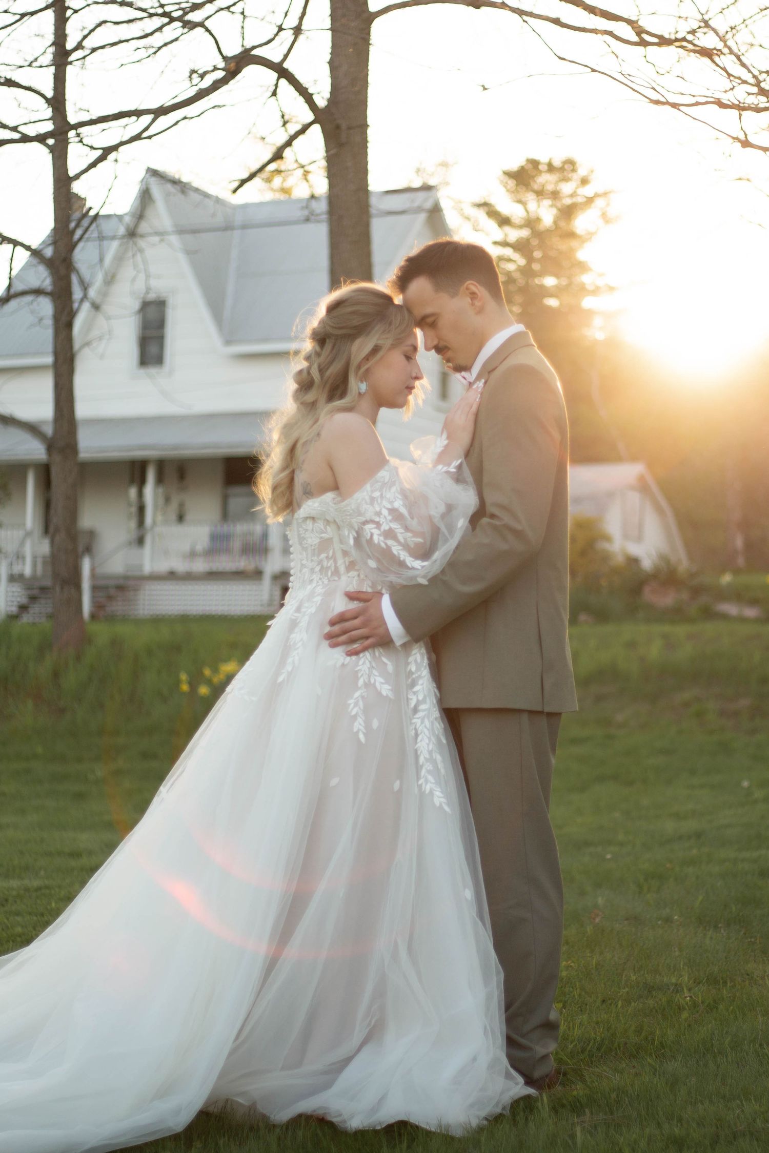Newlyweds embrace in romantic sunset moment on lawn with white farmhouse in background.