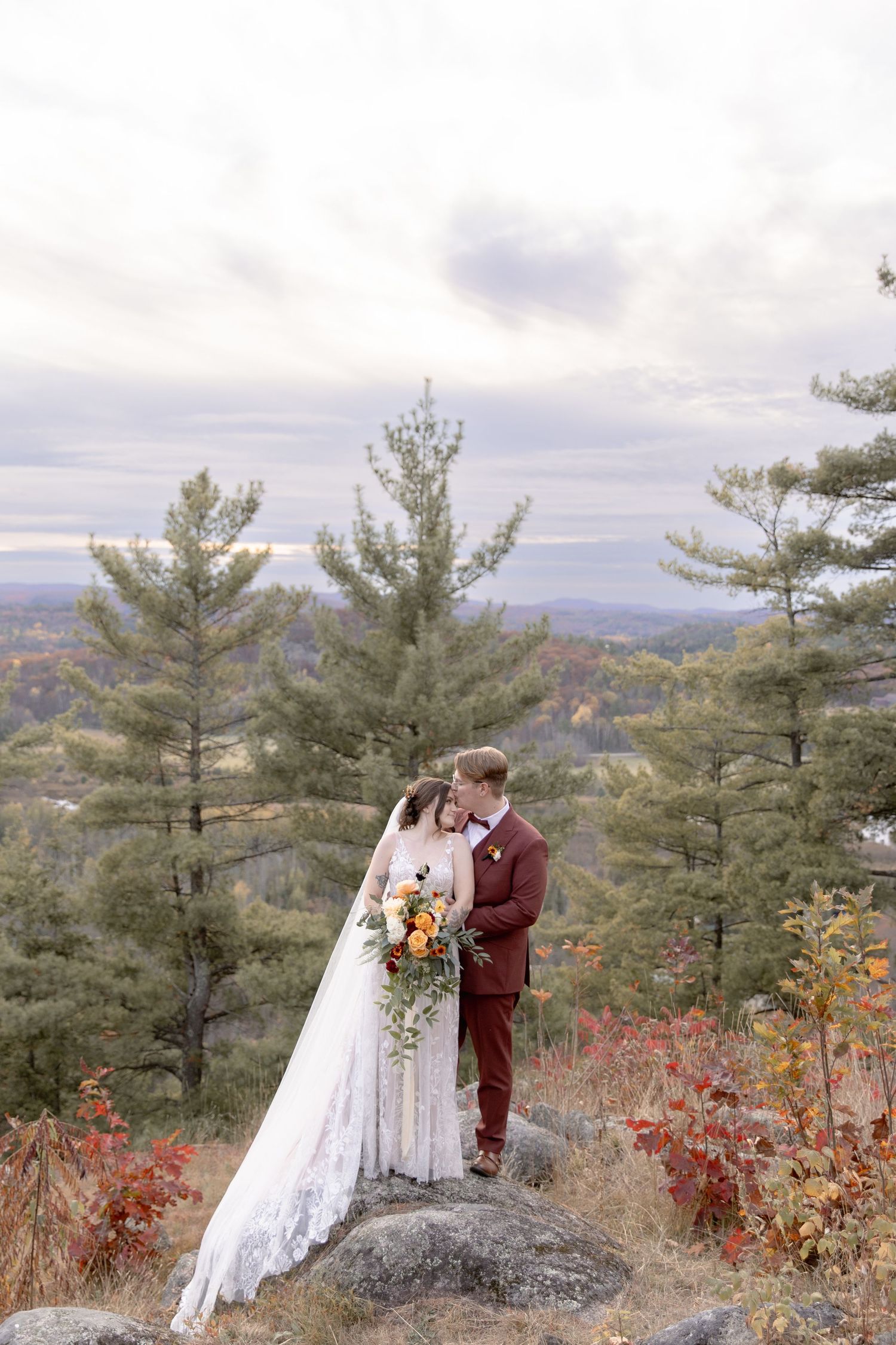A couple in wedding attire stands on a rocky outcrop surrounded by pine trees and autumn foliage with mountain views.