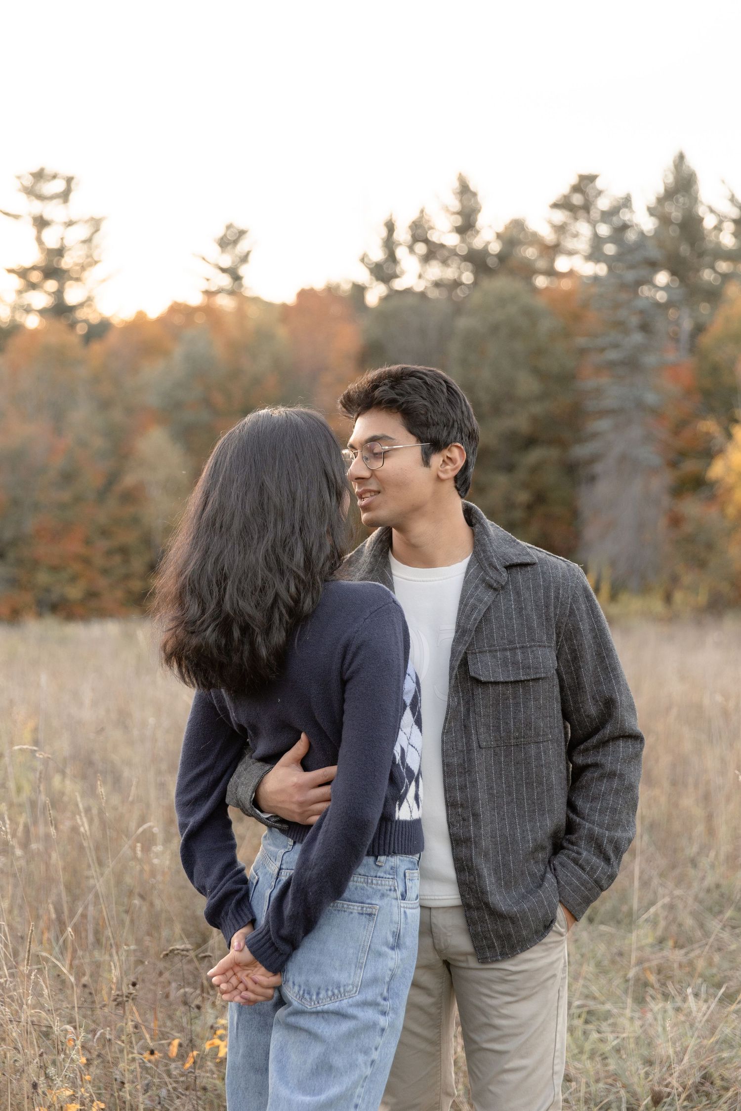 A couple embraces in a golden field at sunset, with one wearing overalls and the other in a plaid blazer.