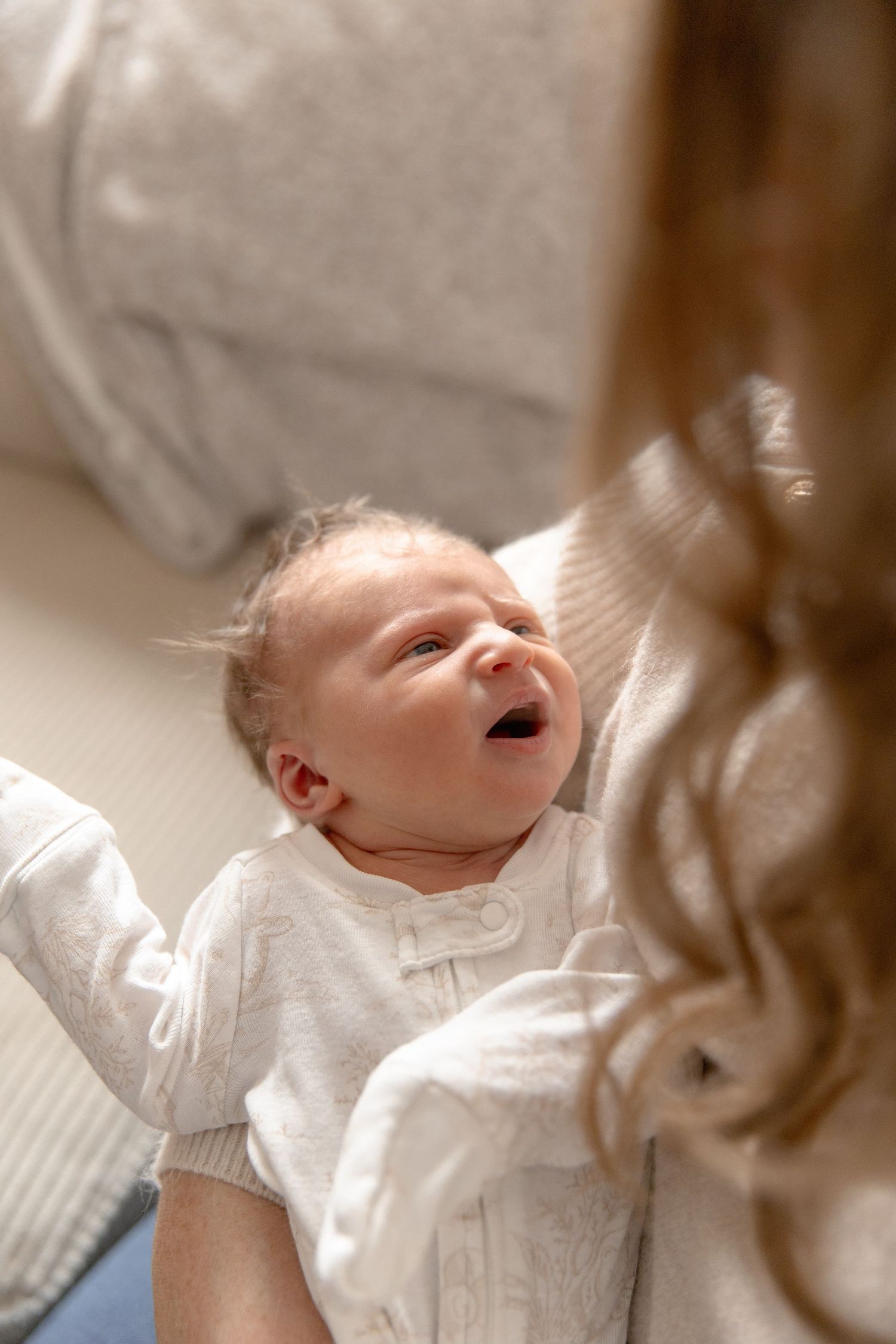 A newborn baby wearing a white christening gown yawns while lying on soft bedding.