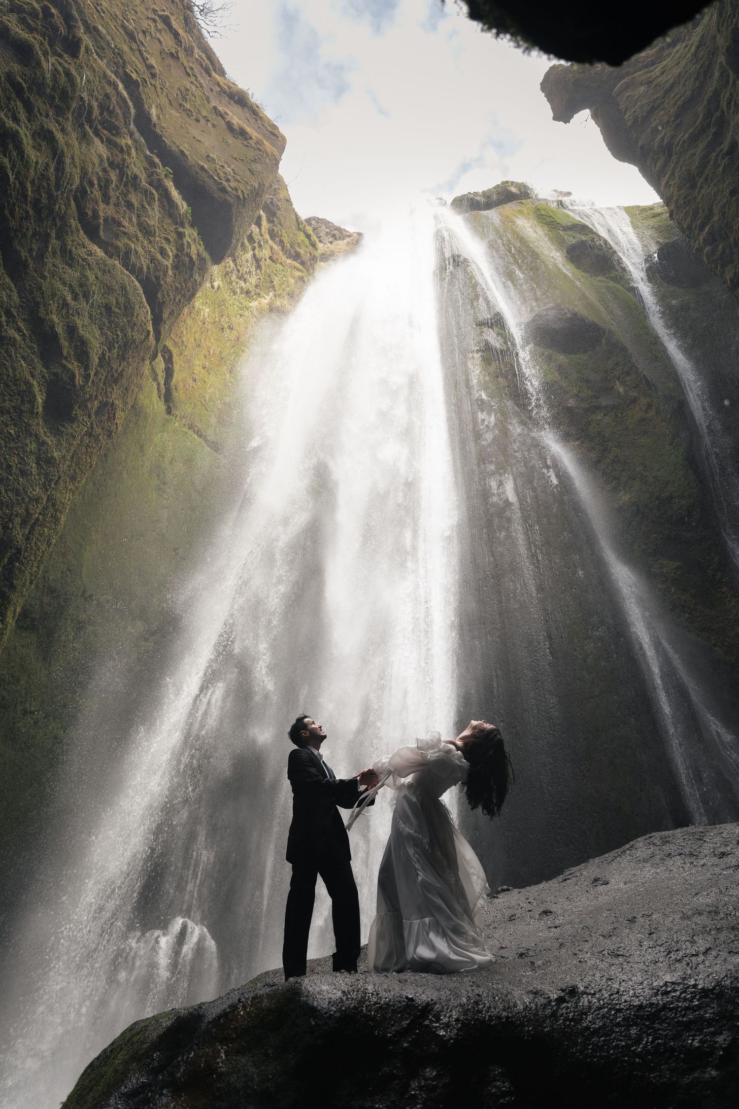 Couple in wedding attire standing before a majestic waterfall cascading down mossy cliffs in Iceland.