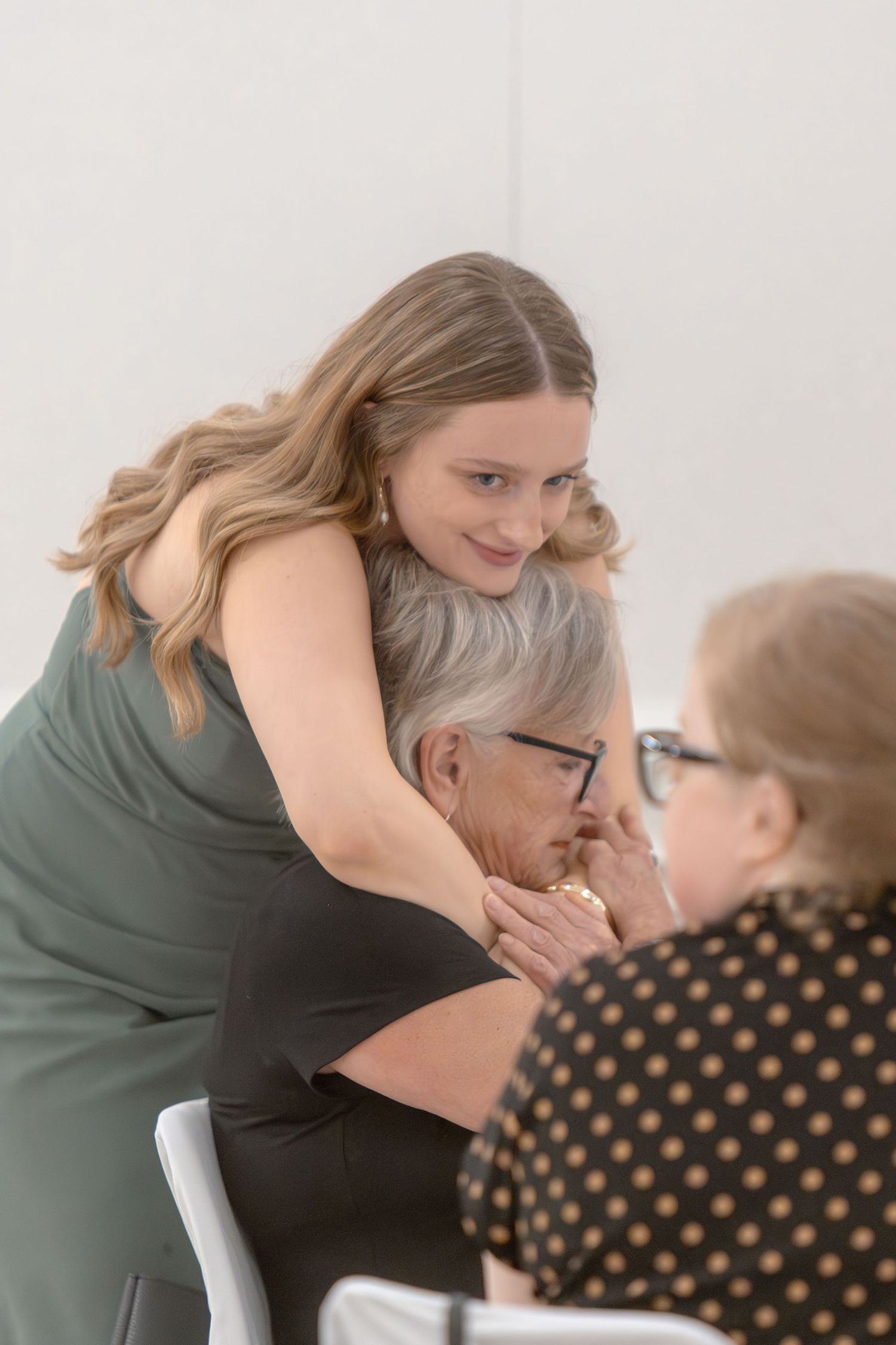 Two people share an emotional embrace in a bright white room while seated in chairs.