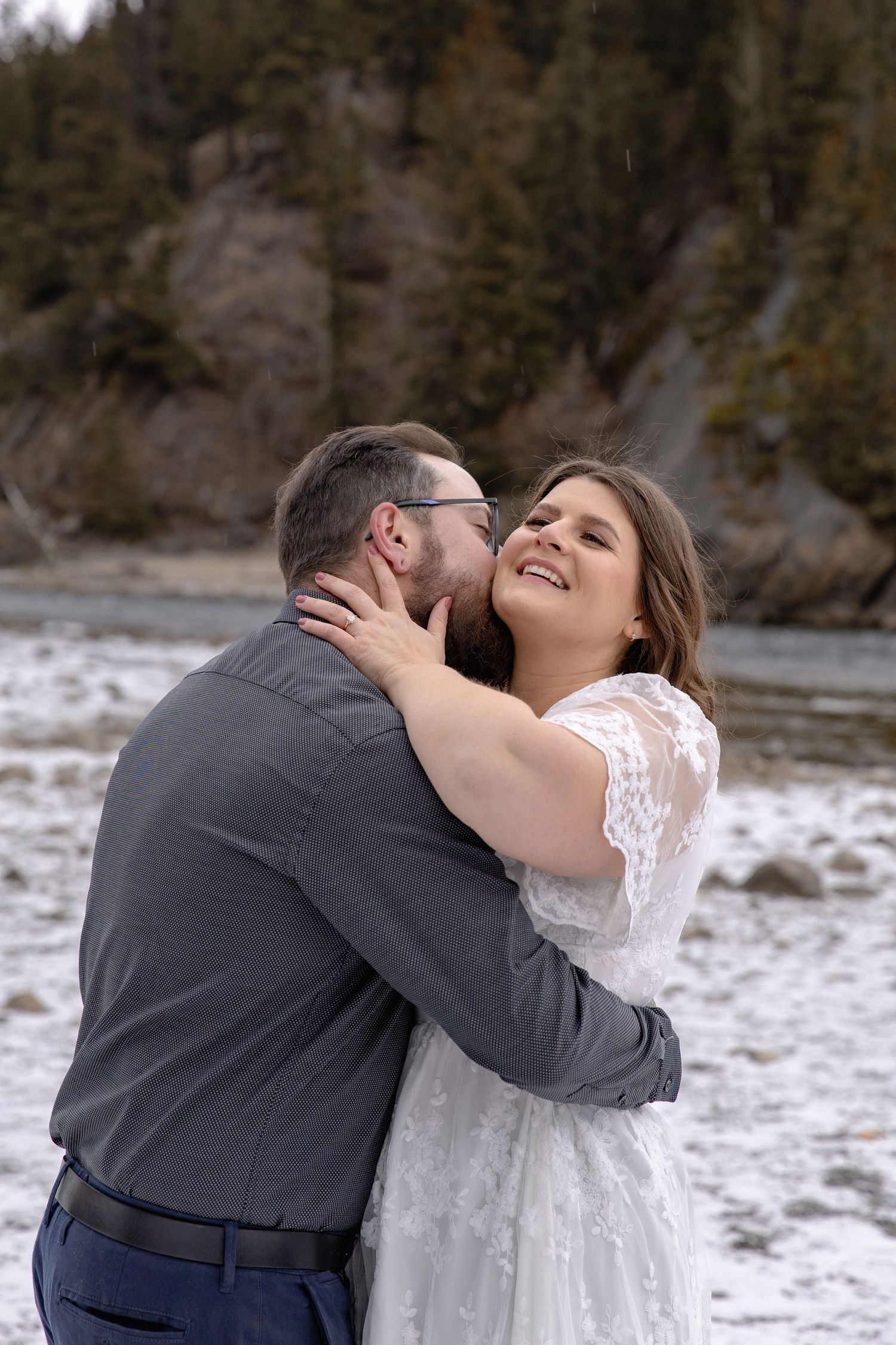 A couple shares a romantic embrace on a snowy riverbank with evergreen trees and rocky cliffs in the background.