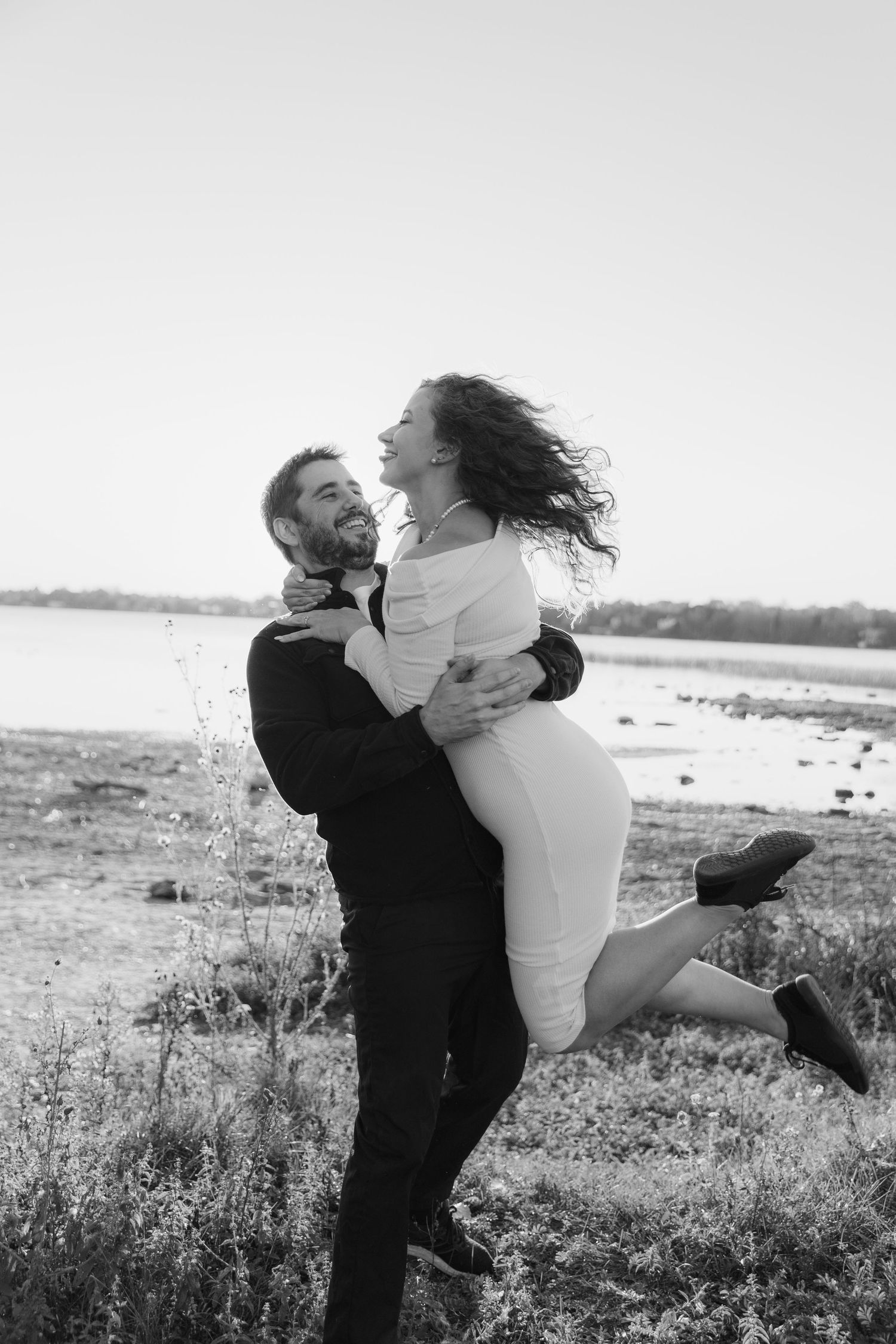 A joyful couple embraces and spins on a beach shoreline in black and white photography.