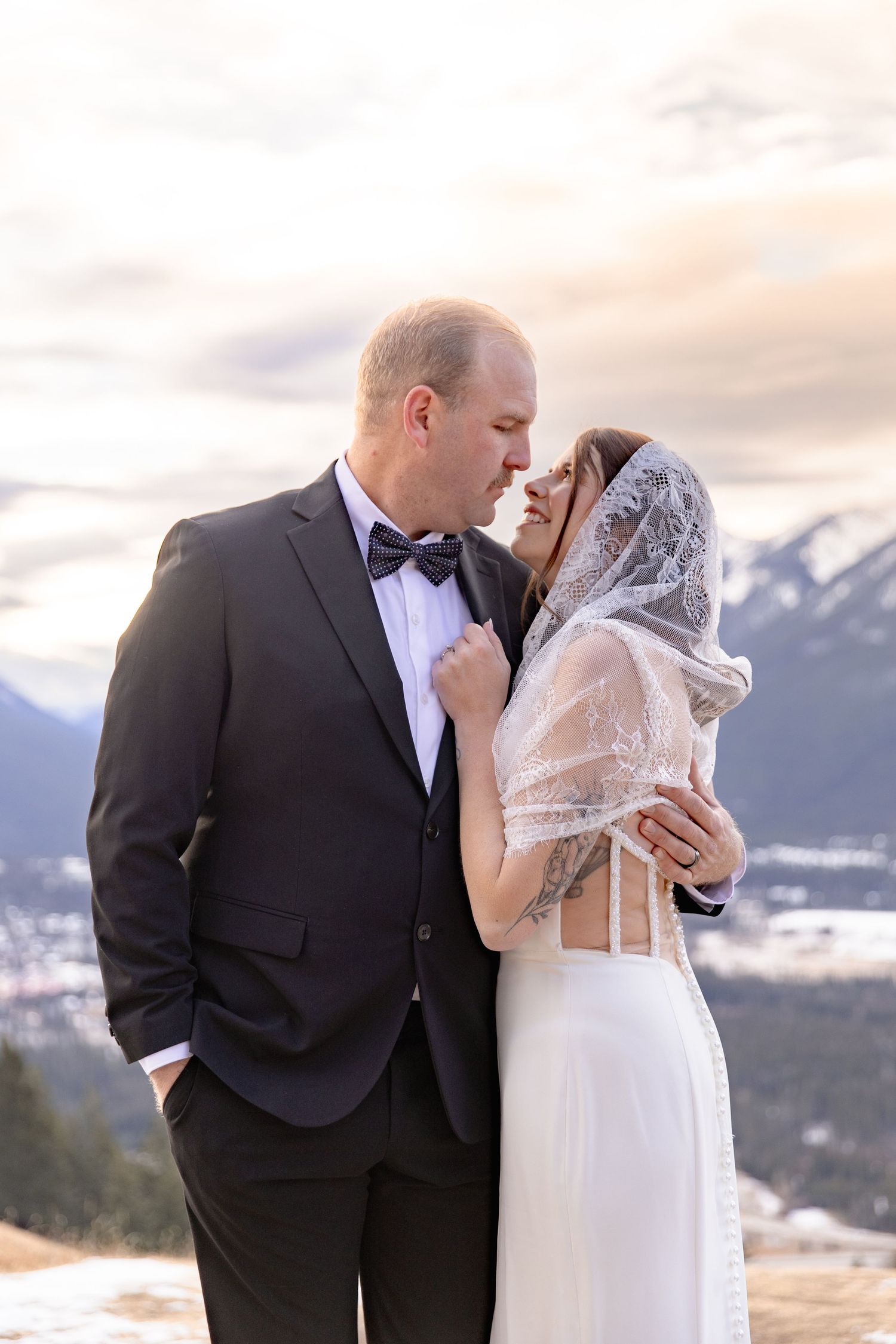 Newlyweds embrace tenderly with snow-capped mountains and warm sunset sky in the background.