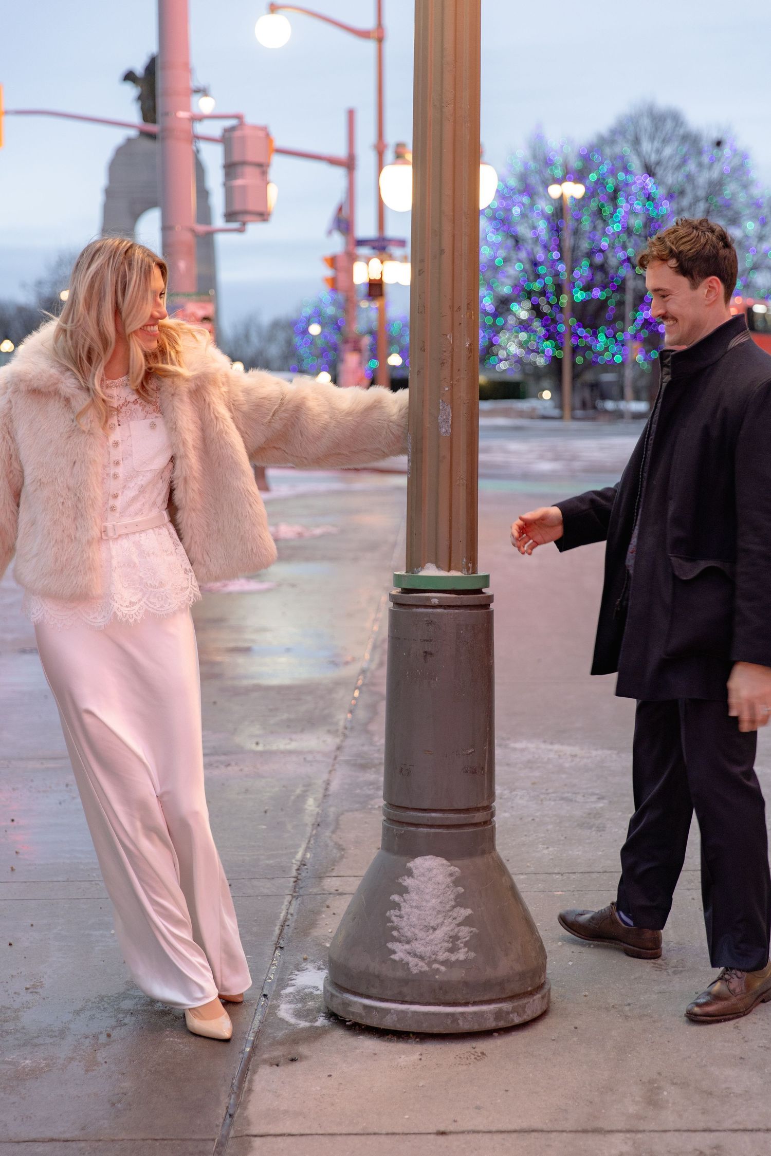 Two people interact playfully on a city sidewalk at dusk, with one wearing a pink furry coat and the other in dark attire.