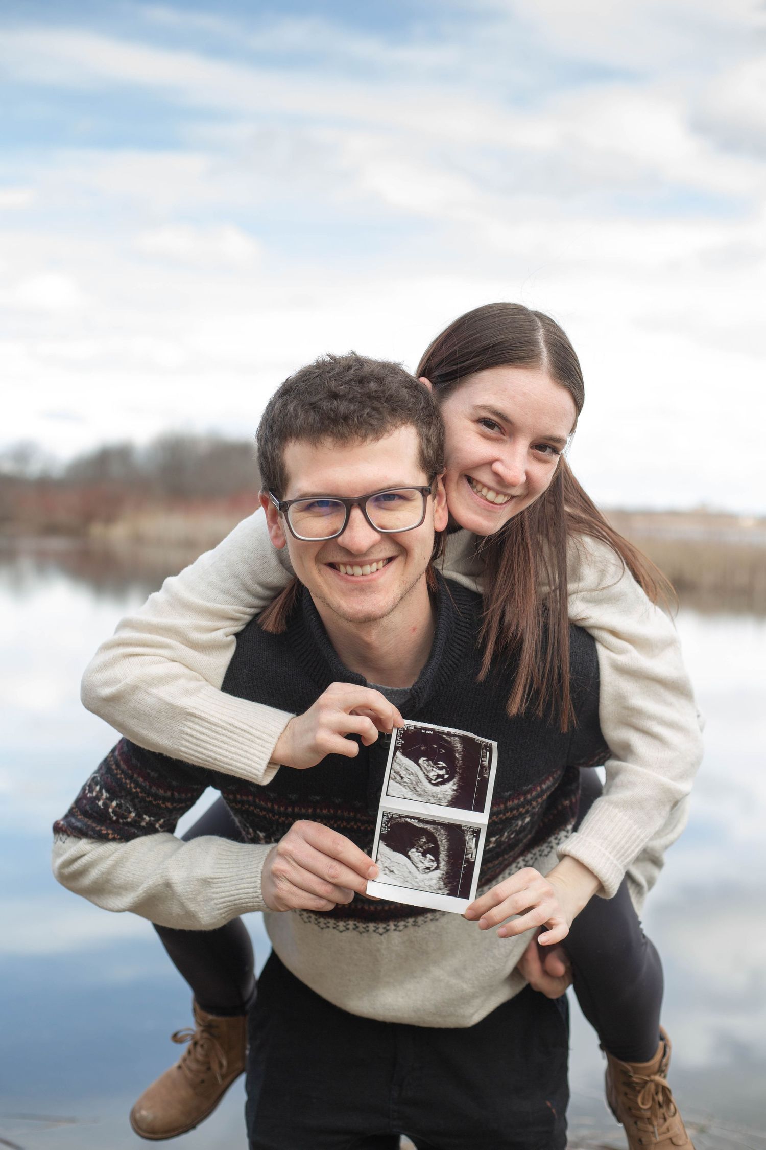 A man gives his girlfriend a piggyback outdoors while holding an ultrasound photo near a lake.