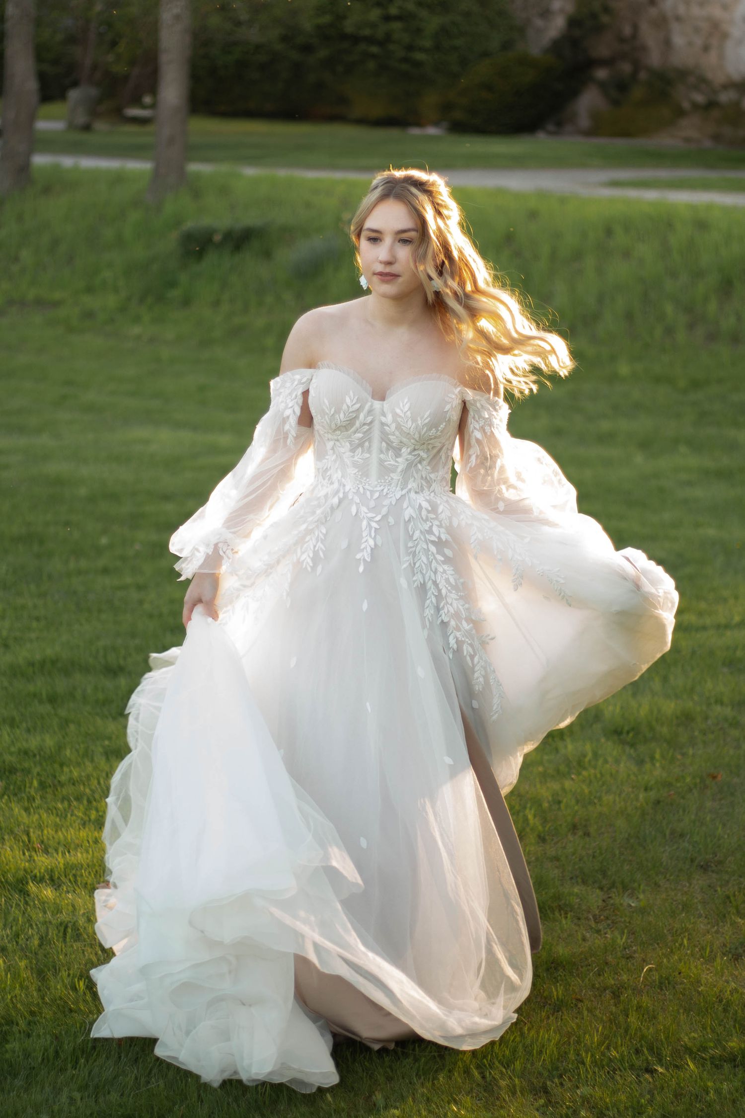 Bride in off-shoulder floral lace gown with billowing skirt poses in sunlit garden setting.