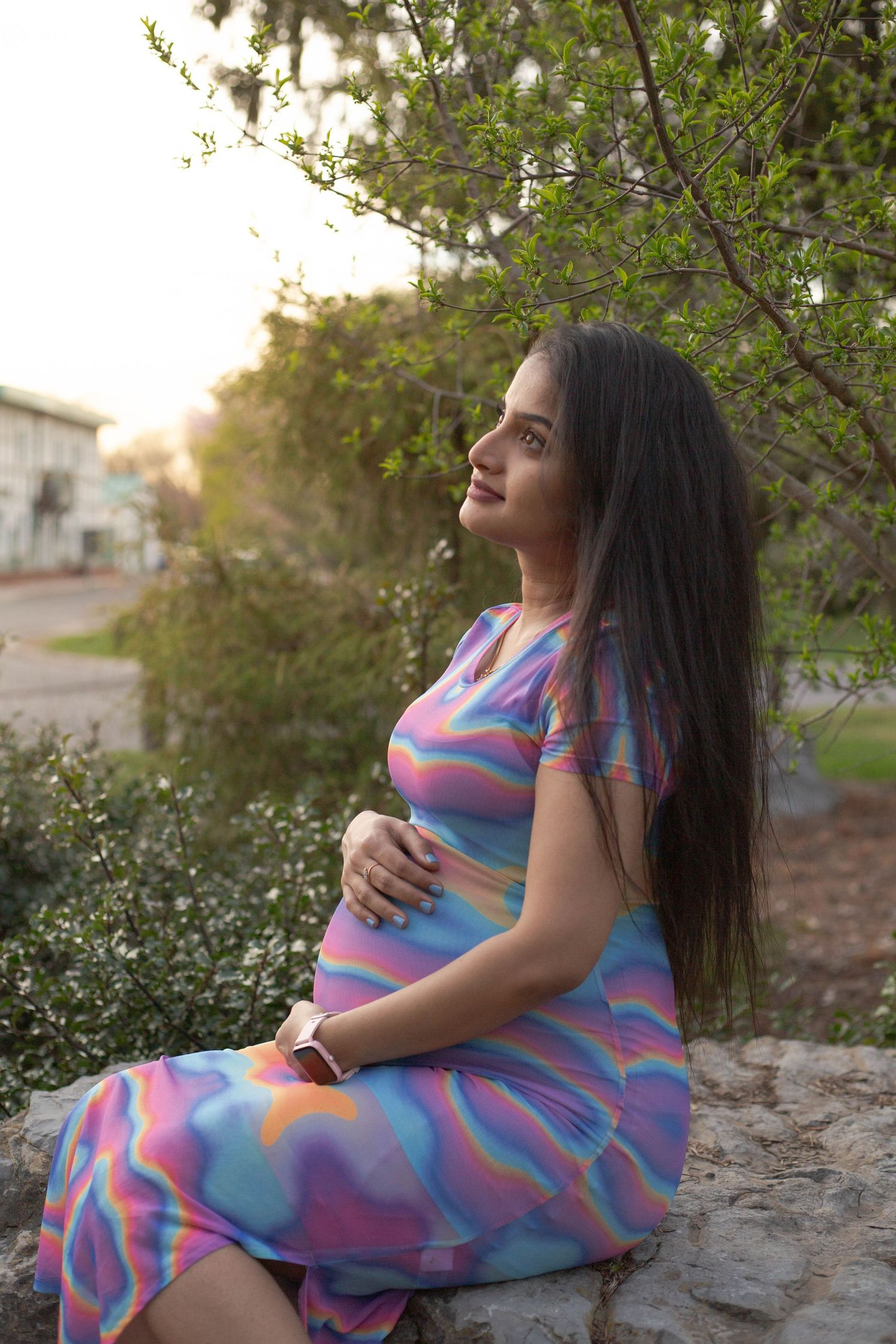 Expectant mother in colorful striped dress sitting on stone wall at sunset among spring blossoms.