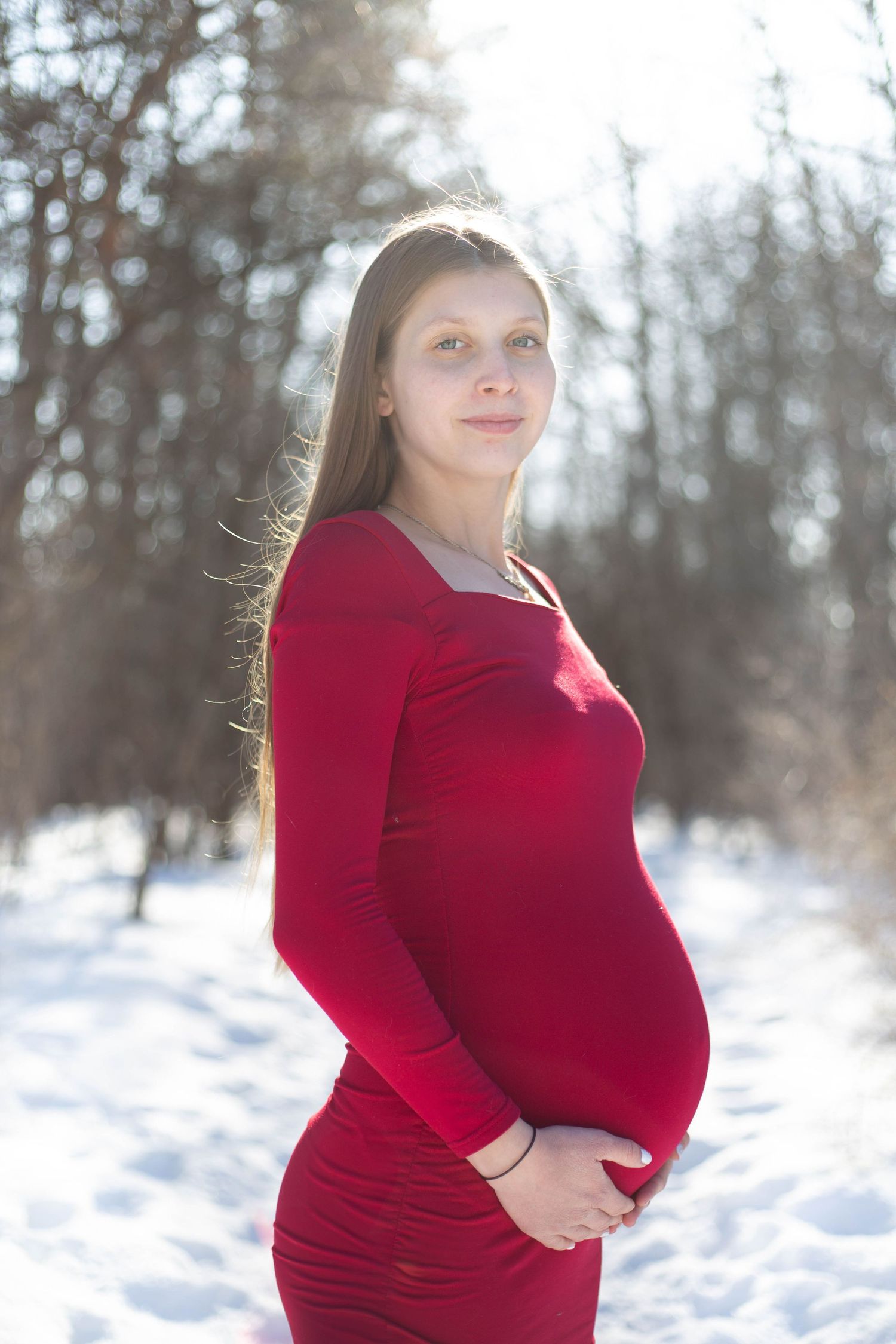 Glowing pregnancy photo with form-fitting red dress in winter forest scene.
