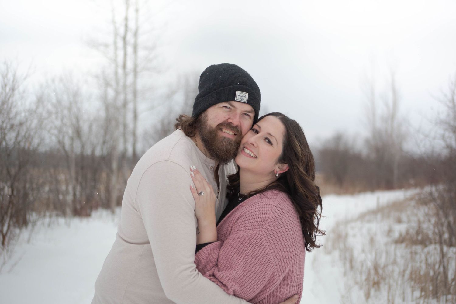 Couple shares tender embrace on snow-covered path with bare trees in background.