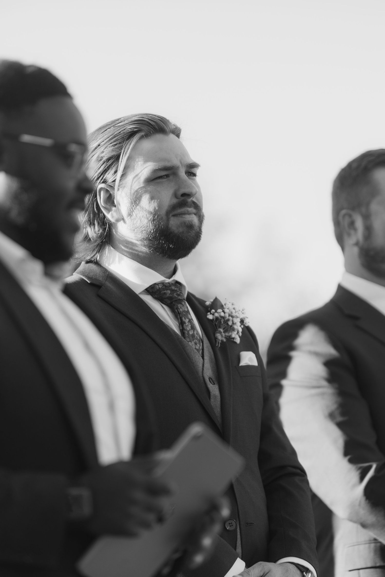 A black and white profile view of groomsmen standing in a line at a formal wedding ceremony.
