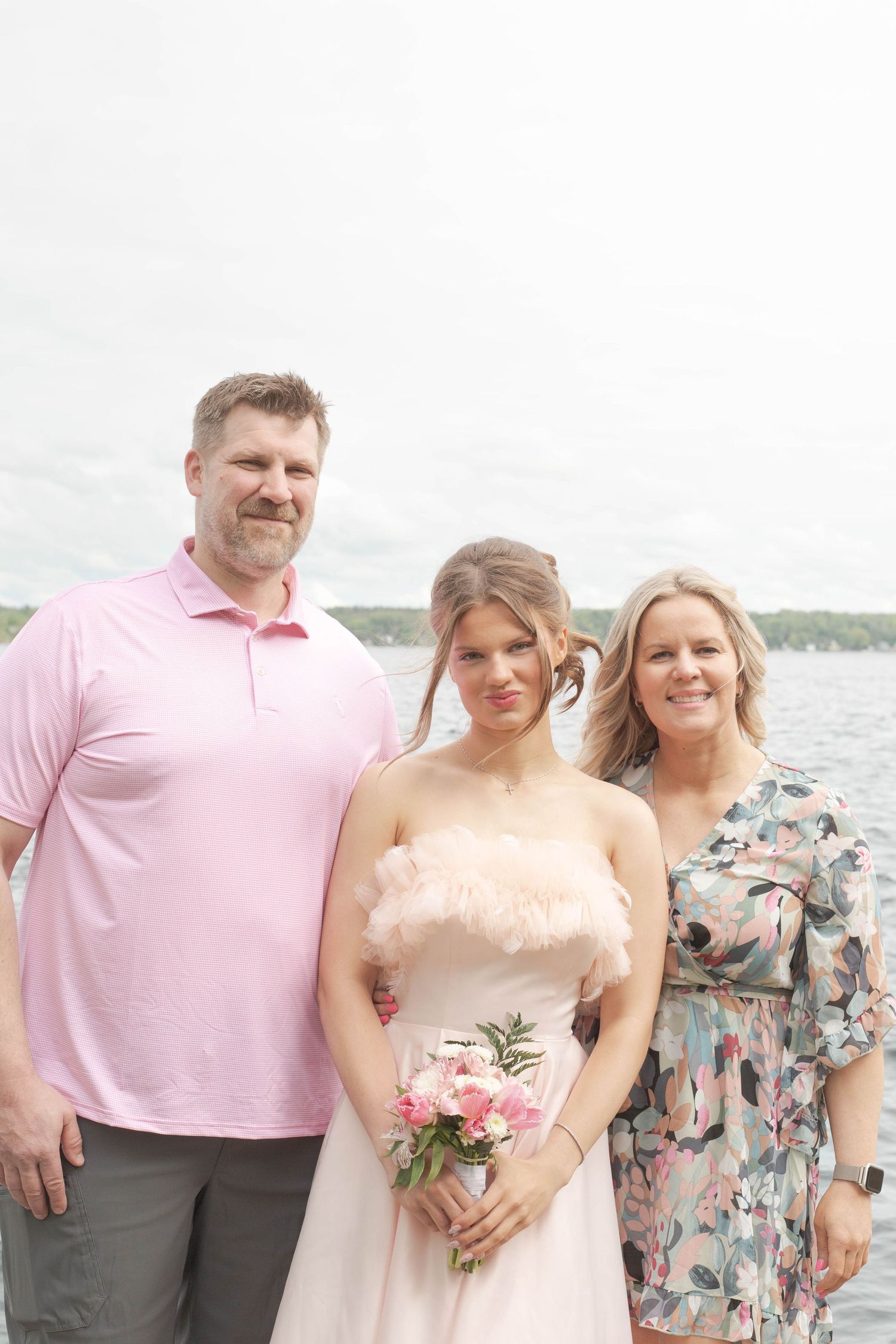 Three people pose together by a lake, person in middle wears a light pink strapless dress holding fresh flowers.