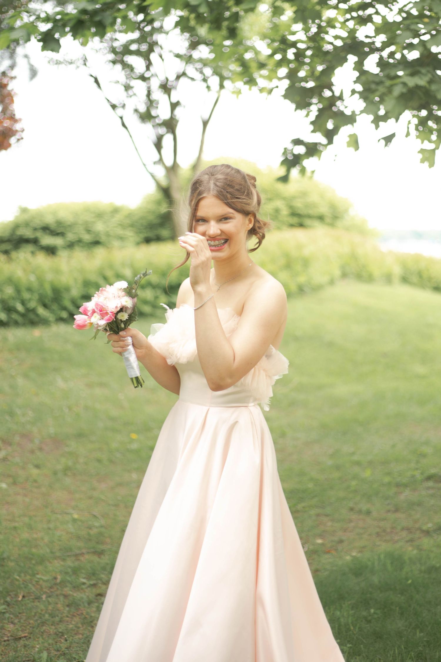 A joyful bride in a blush pink gown holds a small bouquet while standing on a lush green lawn.