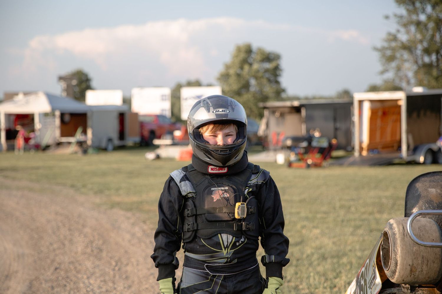A racing driver in full protective gear stands on a dirt track with trailers and vehicles in the background.