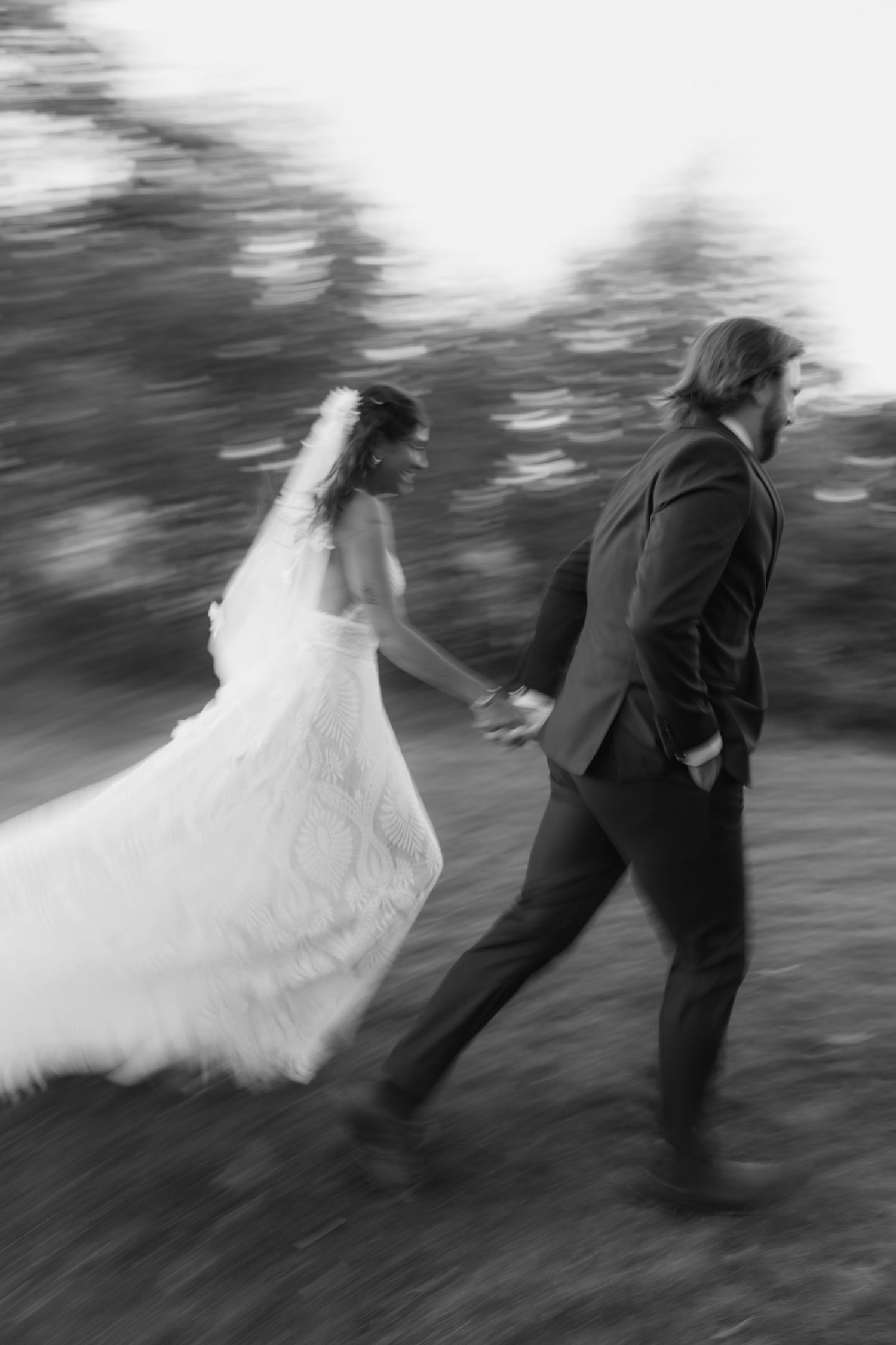 Motion-blurred black and white photo of newlyweds running playfully through a wooded area.
