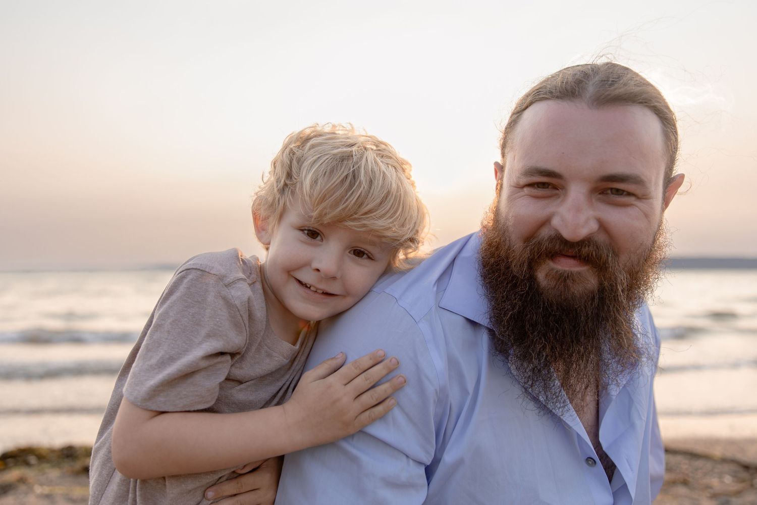 A joyful sunset moment at the beach between an adult with a full beard and a young child wearing gray.