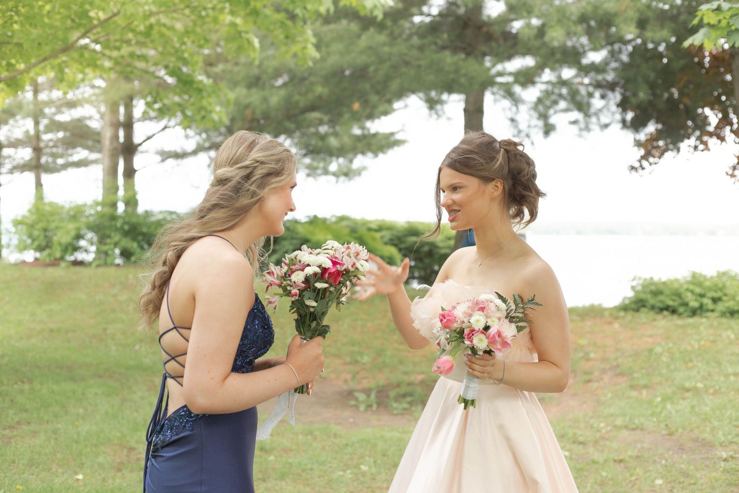 Two people in formal attire share a joyful moment holding pink floral bouquets in an outdoor garden setting by a lake.