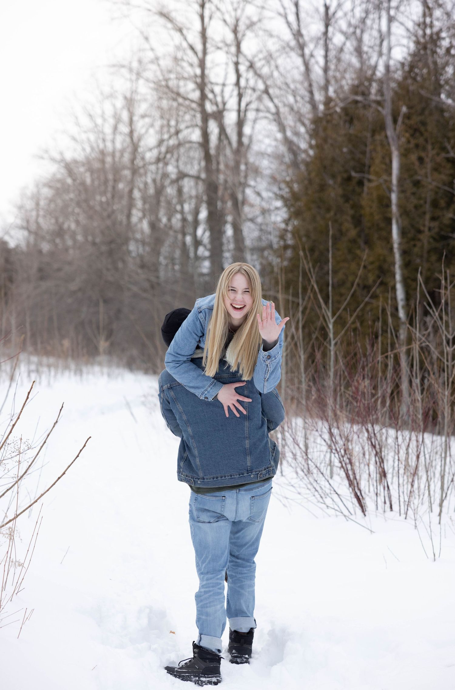 Man in denim jacket and jeans walks away from camera carrying fiancé over his shoulder in winter landscape with bare trees in background.