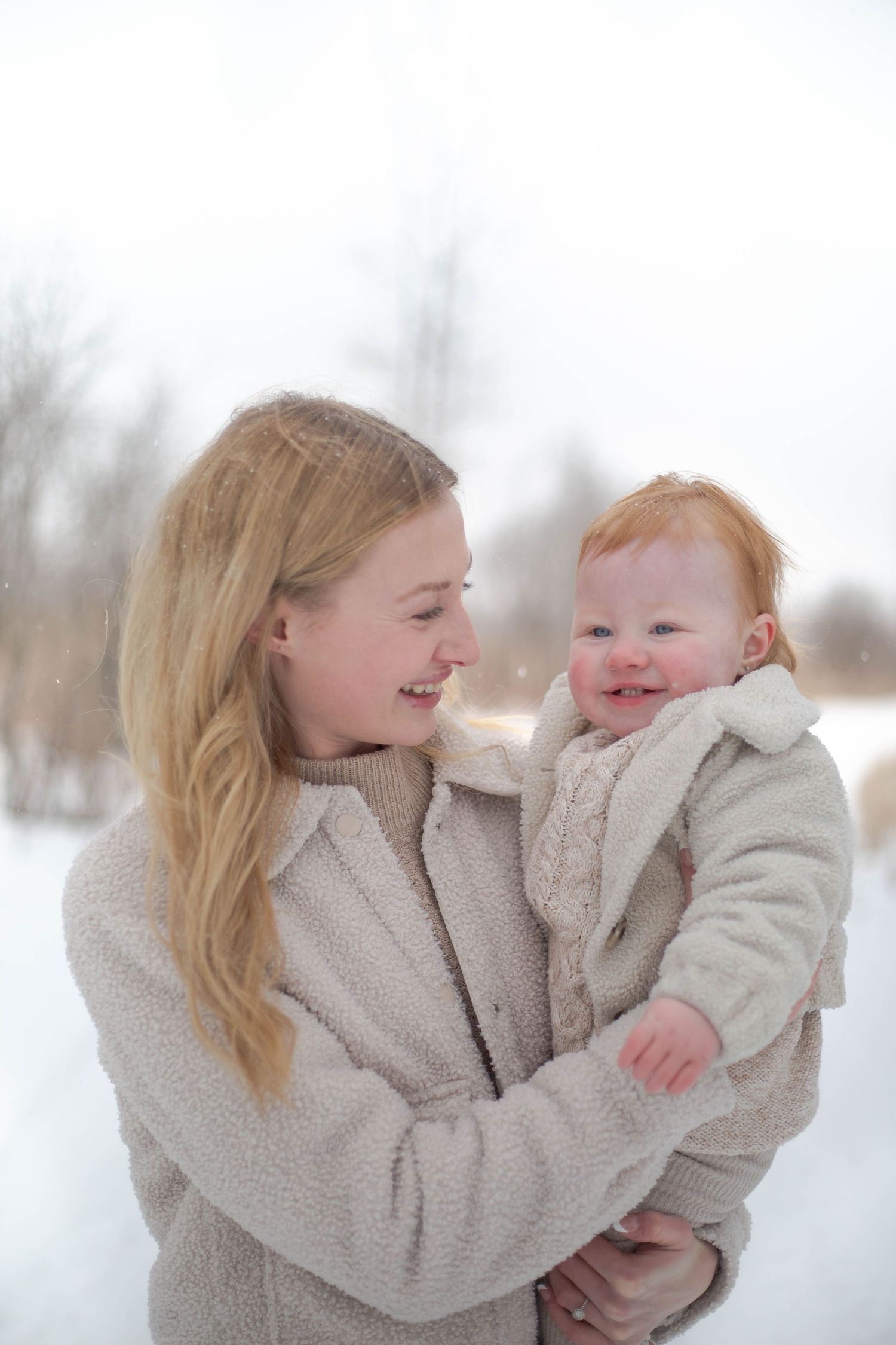 A mom and baby share joyful laughter while embracing in matching cream colored outfits.