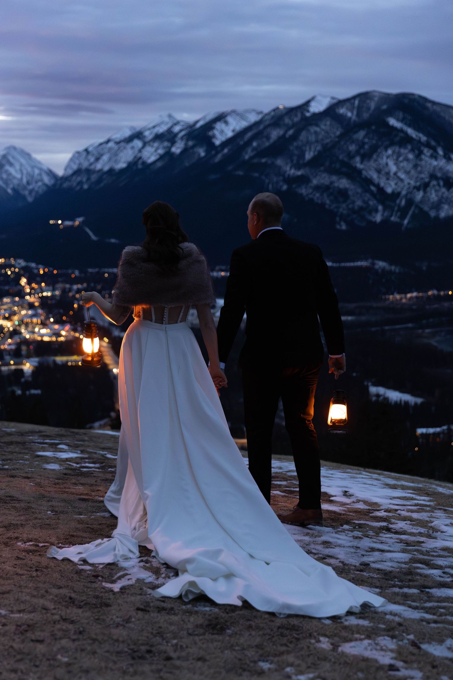 A couple in wedding attire gazes at the twinkling lights of a mountain valley at dusk with lanterns nearby.