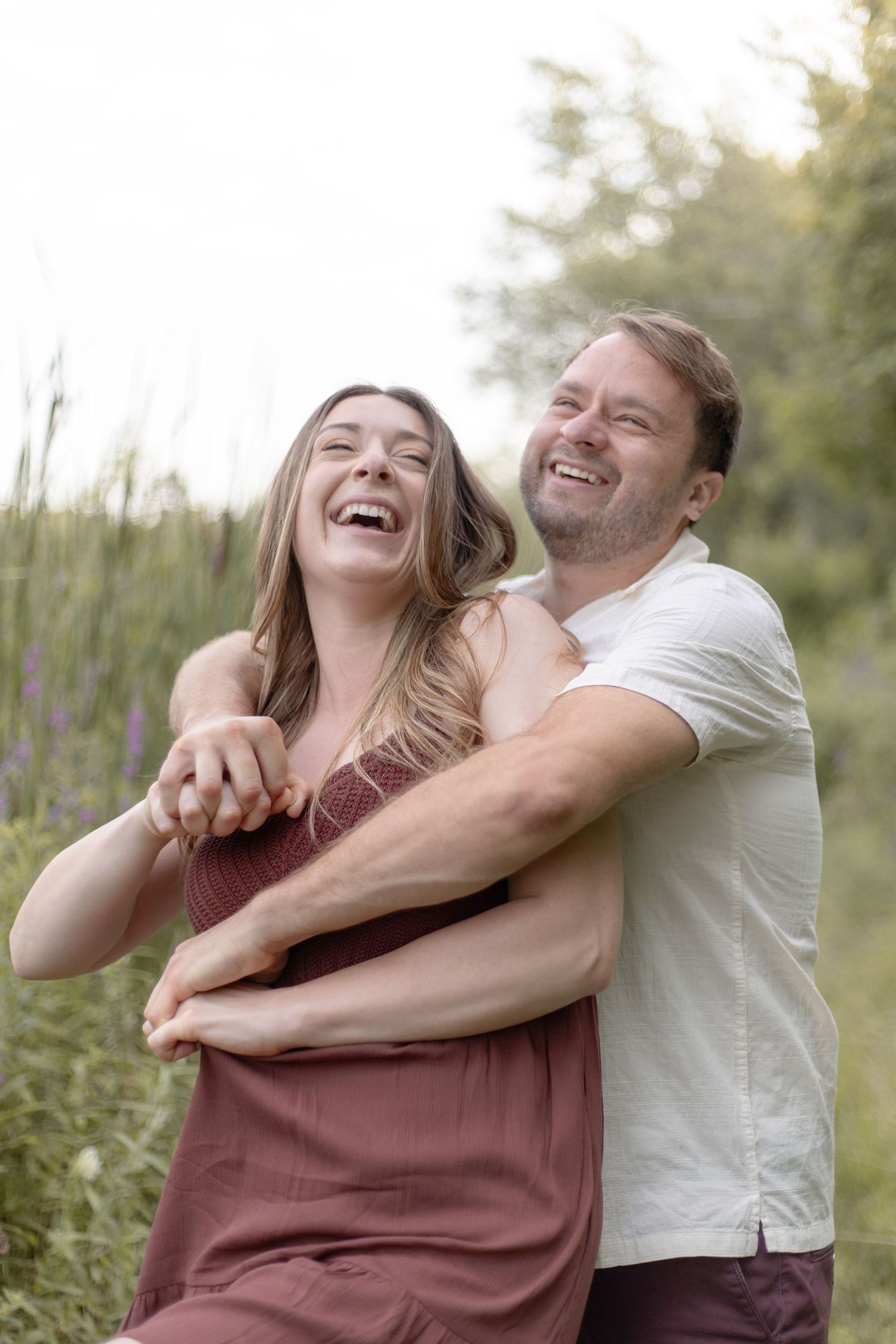 A couple laughs joyfully together in a natural outdoor setting with tall grass and soft lighting.