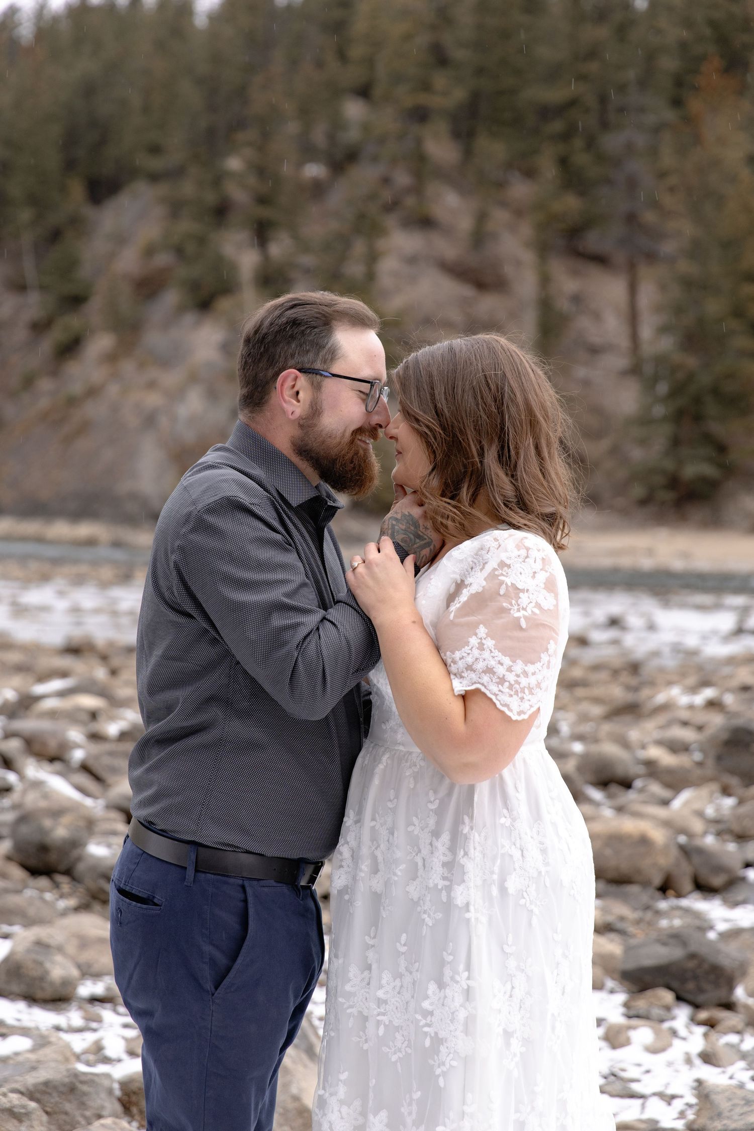 A couple shares a tender kiss in front of a snowy mountain landscape with evergreen trees in the background.