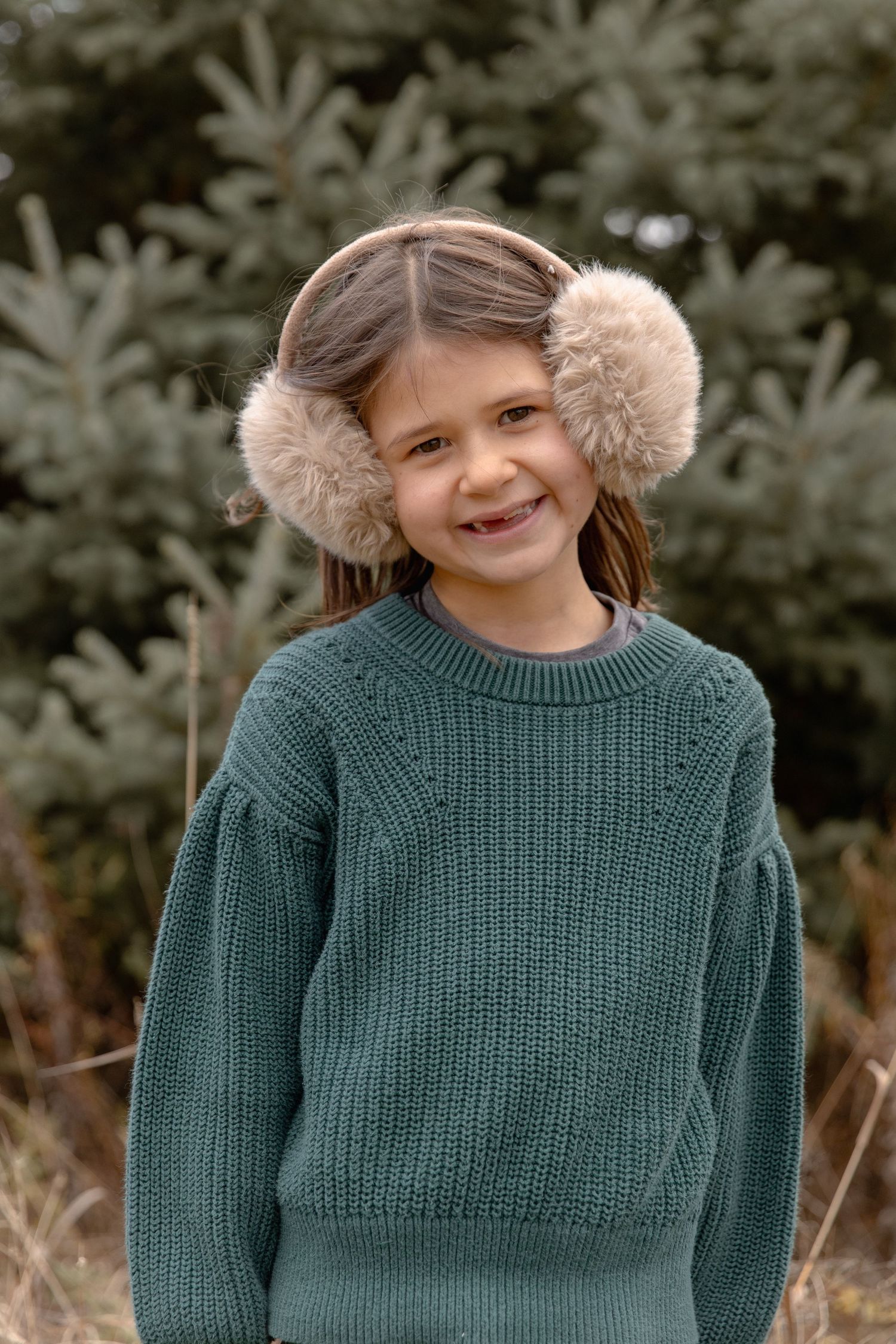 A child wearing a green knit sweater and fuzzy earmuffs poses against a blurred outdoor background.
