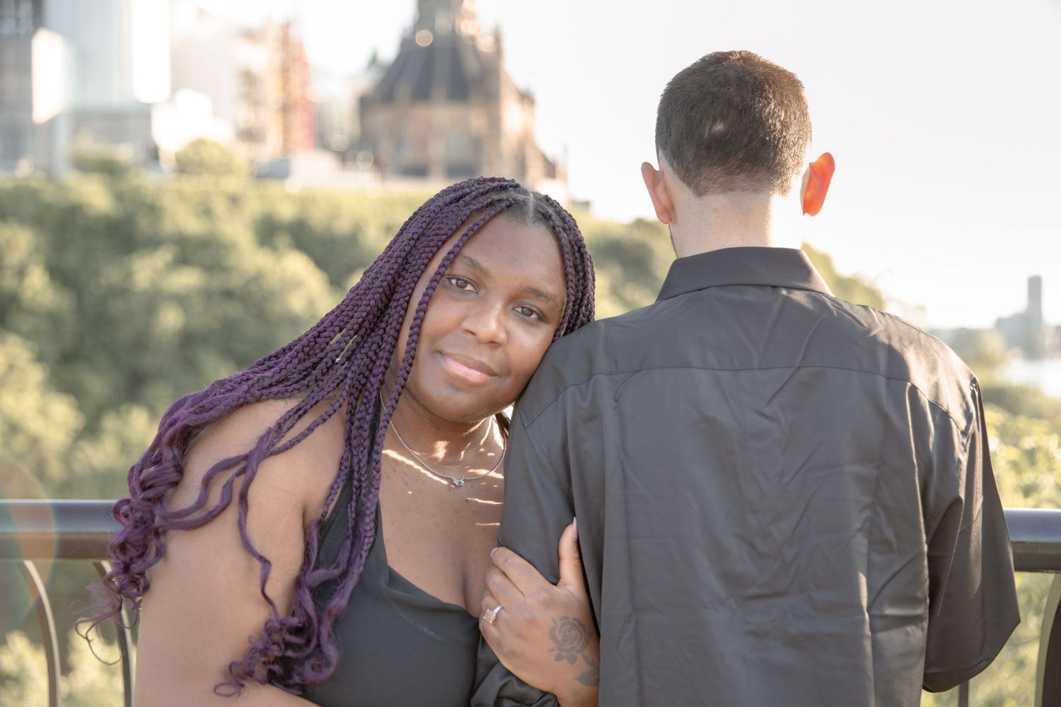 A couple stands together outdoors during golden hour with city buildings visible in the background.