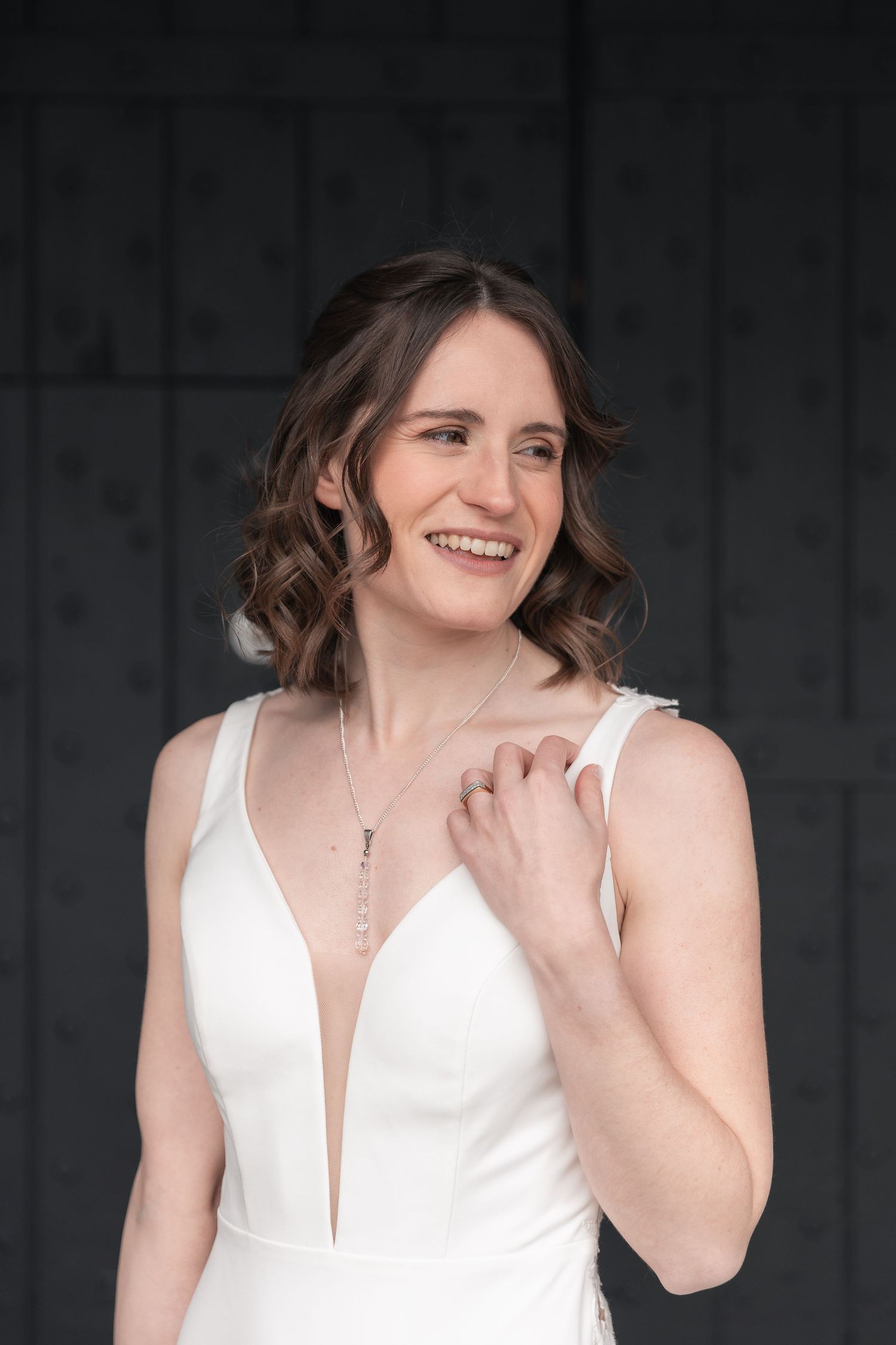 Smiling bride in white deep-V gown with delicate necklace, posing against dark textured wall.