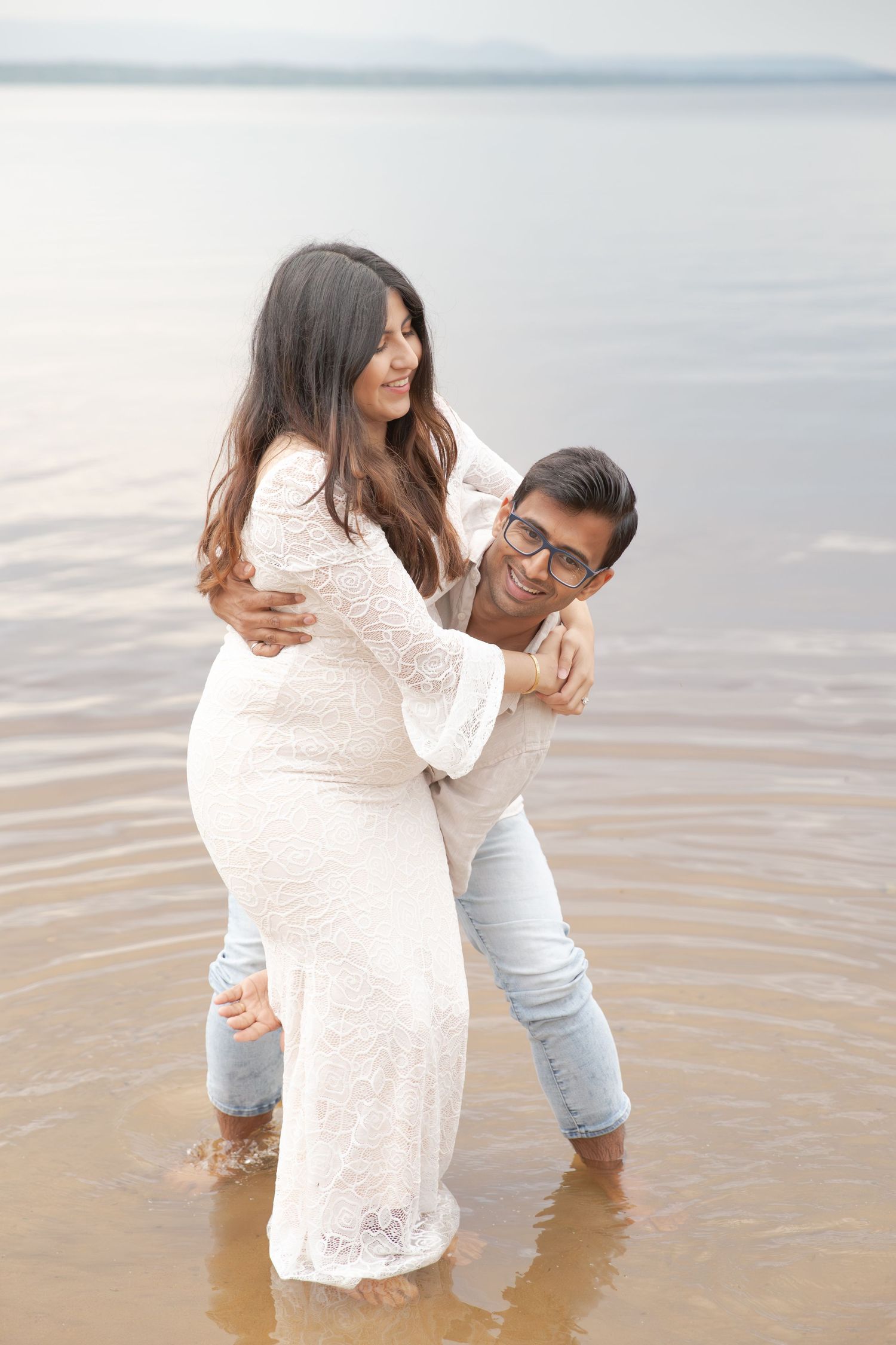 A couple shares a romantic moment in shallow water, with her wearing a white lace dress and him in casual attire.