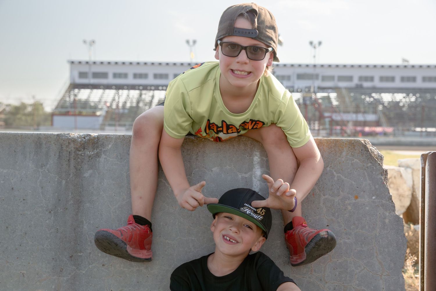 Two people wearing baseball caps crouch on a concrete wall with stadium seating visible in the background.